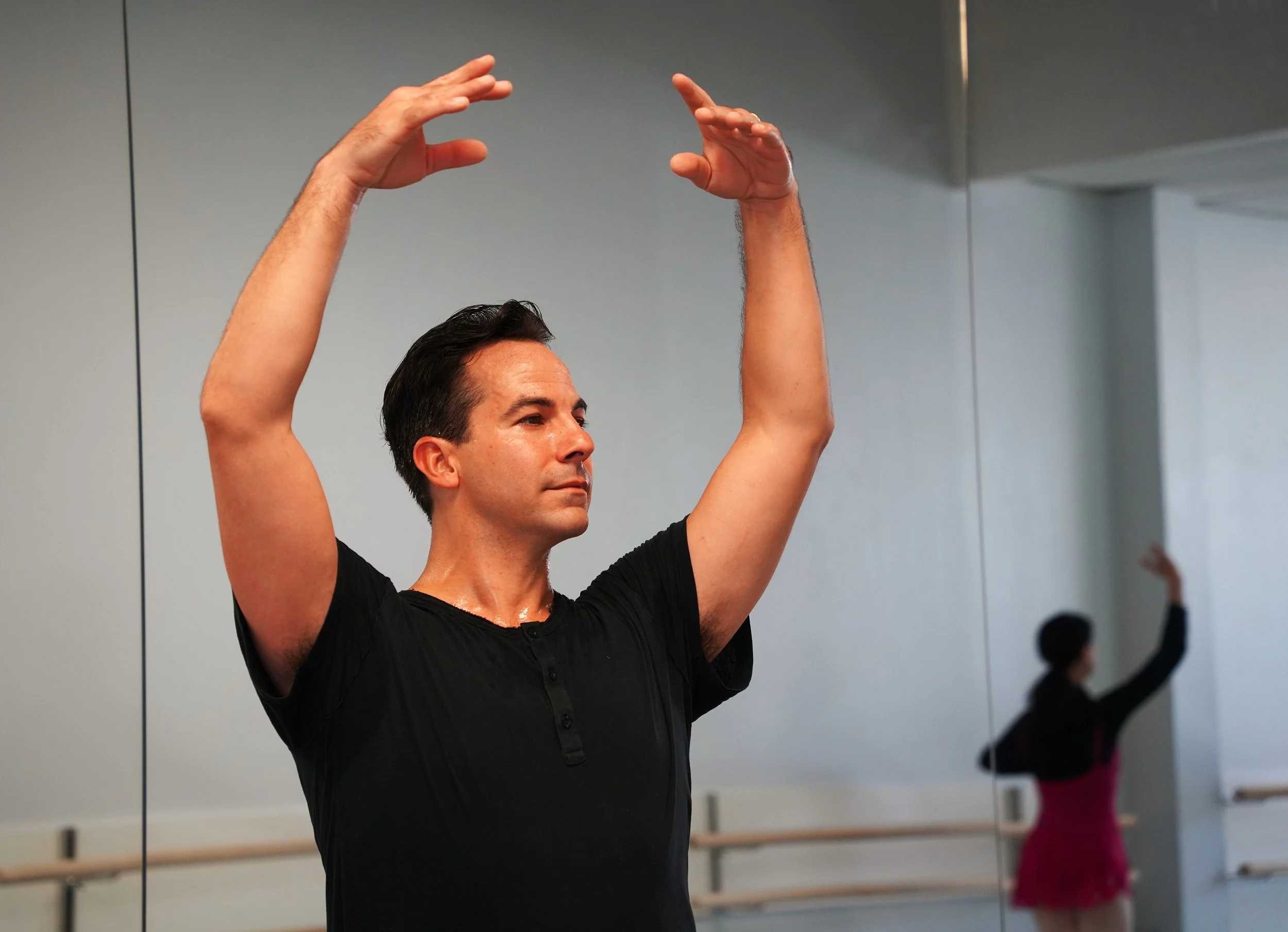 A man with dark hair in a ballet studio, raising his arms in a ballet pose, wearing a black shirt. Mirror reflections show other dancers in the background.