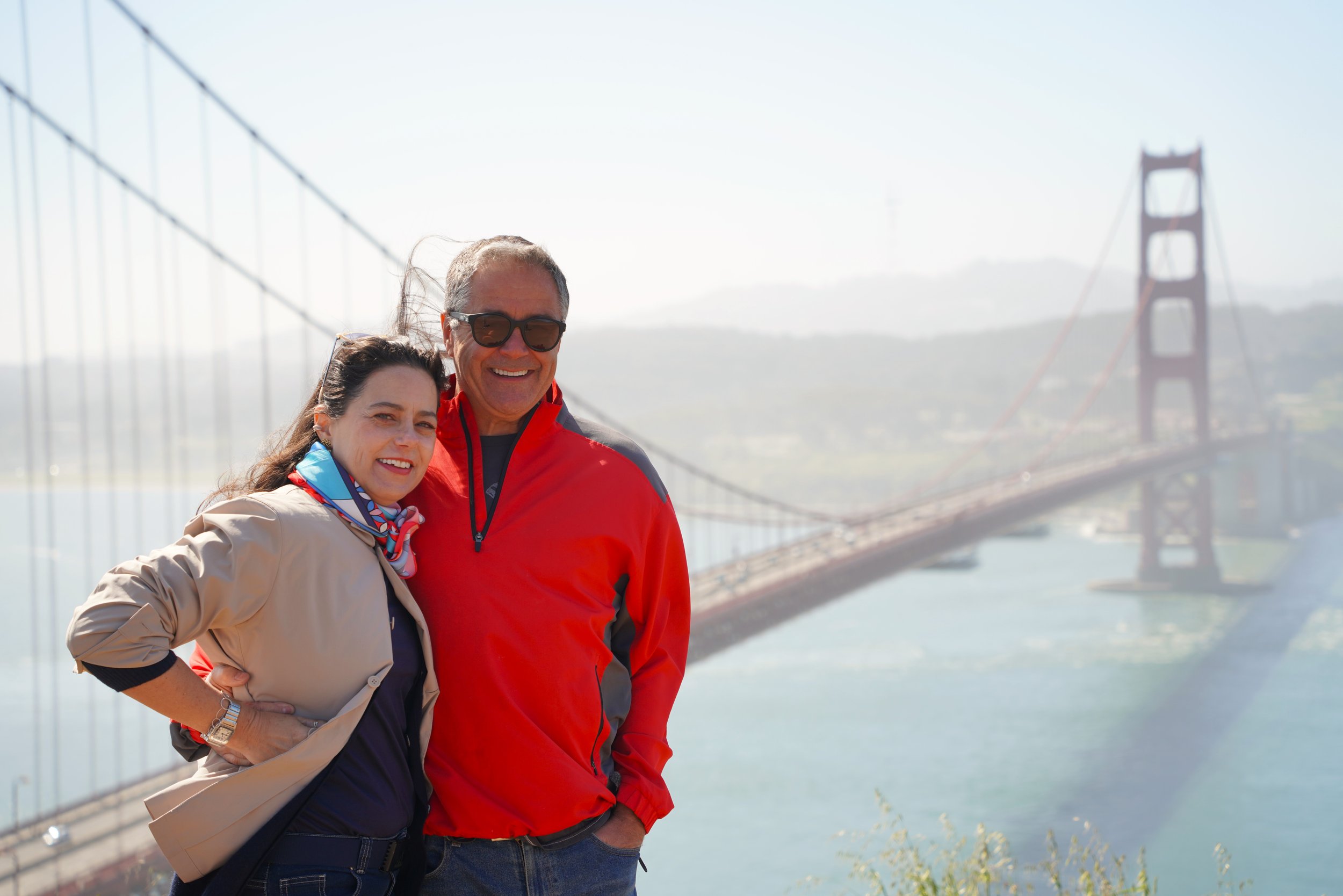 Two people standing together near the Golden Gate Bridge in San Francisco, smiling at the camera, with foggy skies and the bridge's towers in the background.