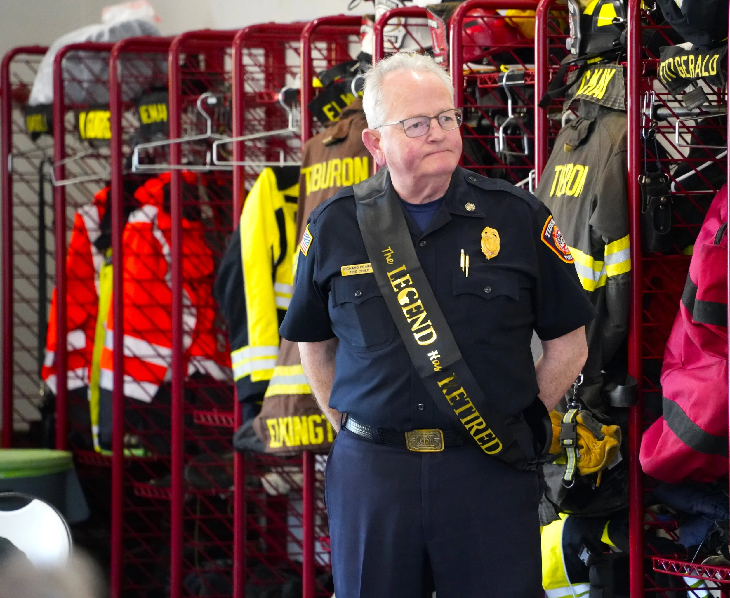 A fire chief wearing glasses and a sash with the words "The Legend" stands in front of a red shelving unit with firefighter gear, including helmets, jackets, and tools.
