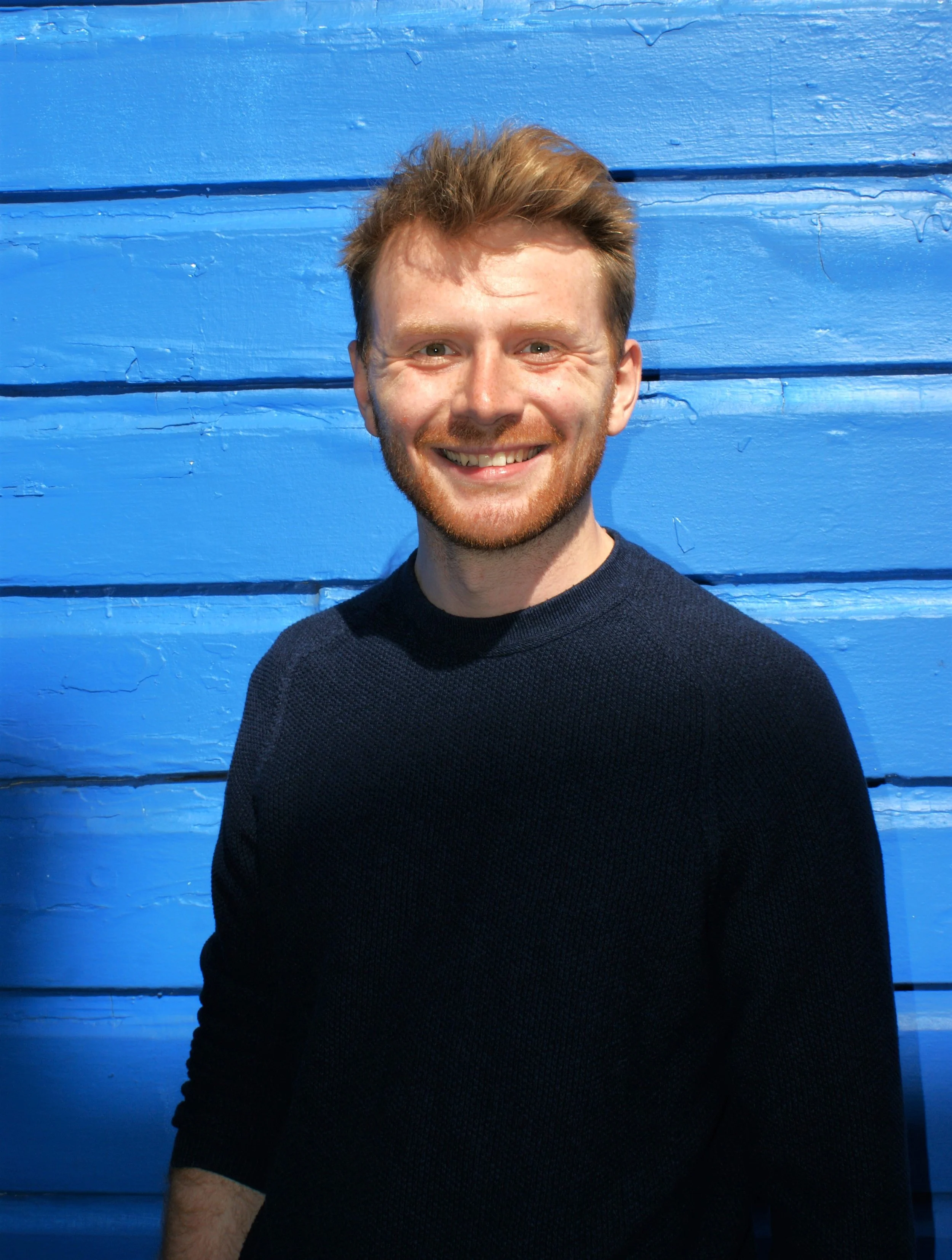 A young man with light brown hair and a beard, smiling, in front of a blue wooden wall.
