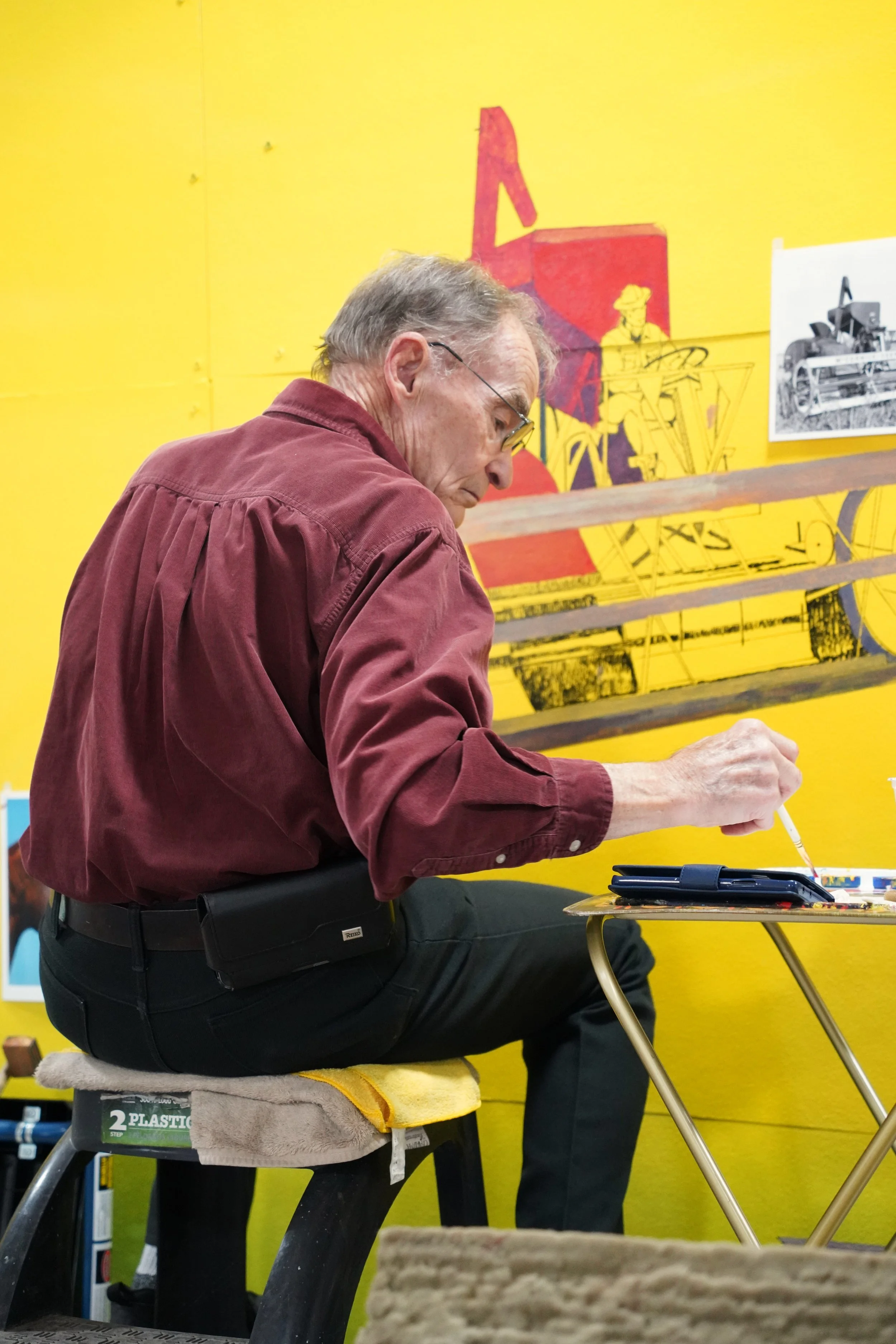 Older man wearing glasses and a red shirt sitting on a stool and working on a project at a table with art supplies, with a yellow background and artwork on the wall.