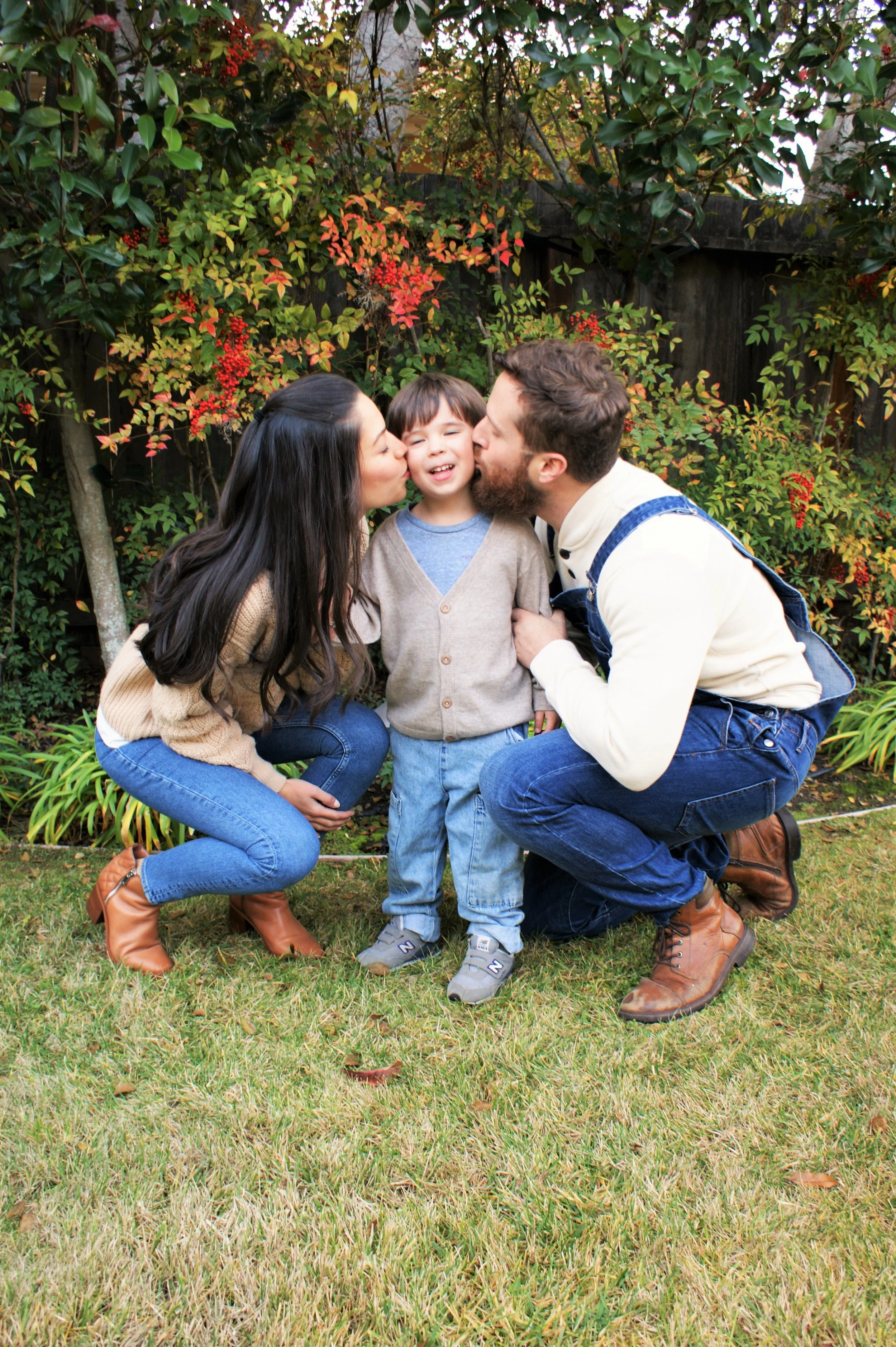 A family of three, a woman, a man, and a young boy, standing outdoors in a garden with colorful autumn foliage in the background. The woman and man are kissing the boy on the cheeks while he smiles happily.