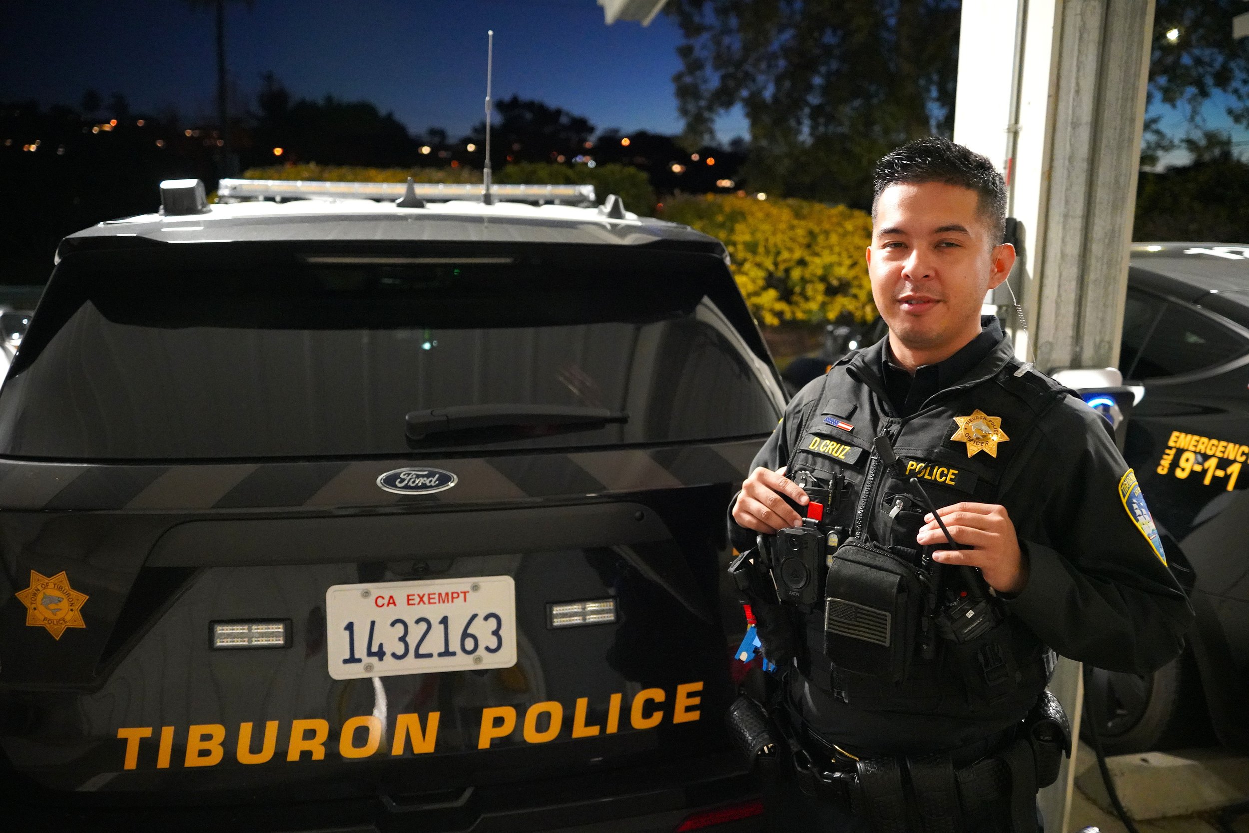 A Tiburon Police officer standing in front of a police vehicle outdoors during night time, holding a device with a mounted camera, wearing a black uniform with police badges and patches, with trees and a dark sky in the background.