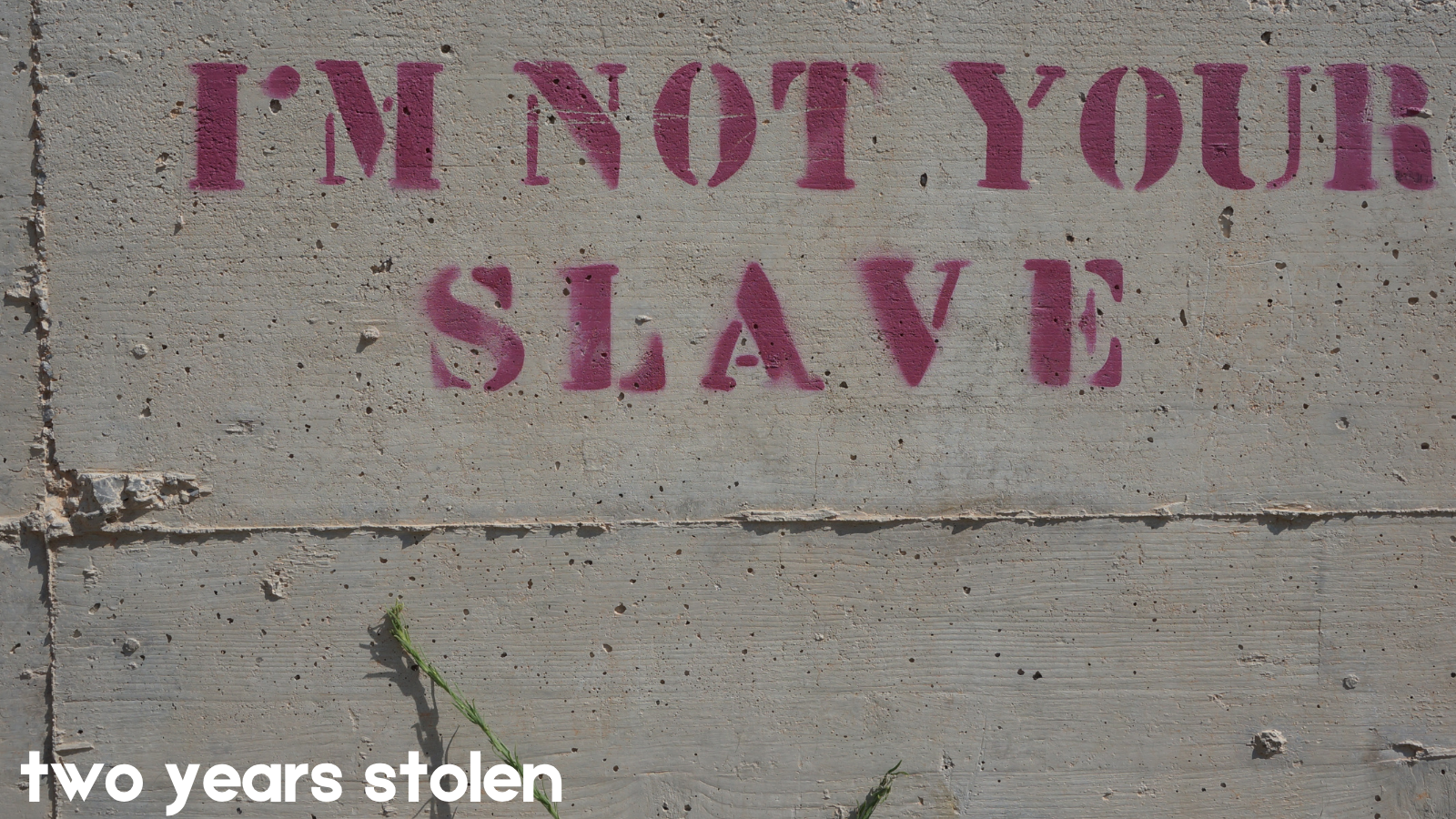 cement wall background with painted stenciled words "I'm Not You Slave"; white overlay letters at bottom left "two years stolen"