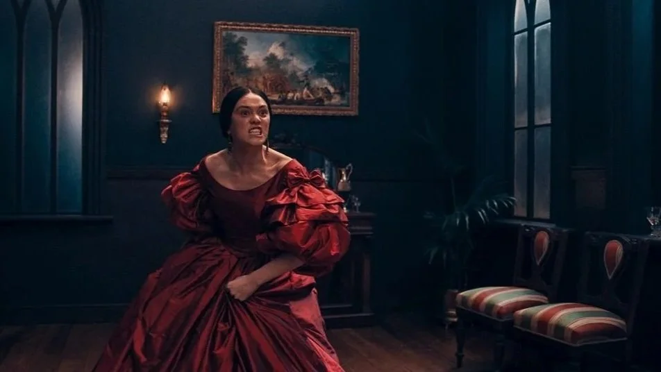 a woman in a Victorian red dress seems angry. Room is a dark parlor with a fireplace and two chairs, a painting hanging above the mantle. Still from Marama.