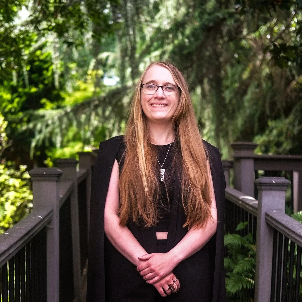Portrait of a woman with long blonde hair, glasses, layered necklaces, and earrings, standing outdoors near a wooden structure with green trees in the background.