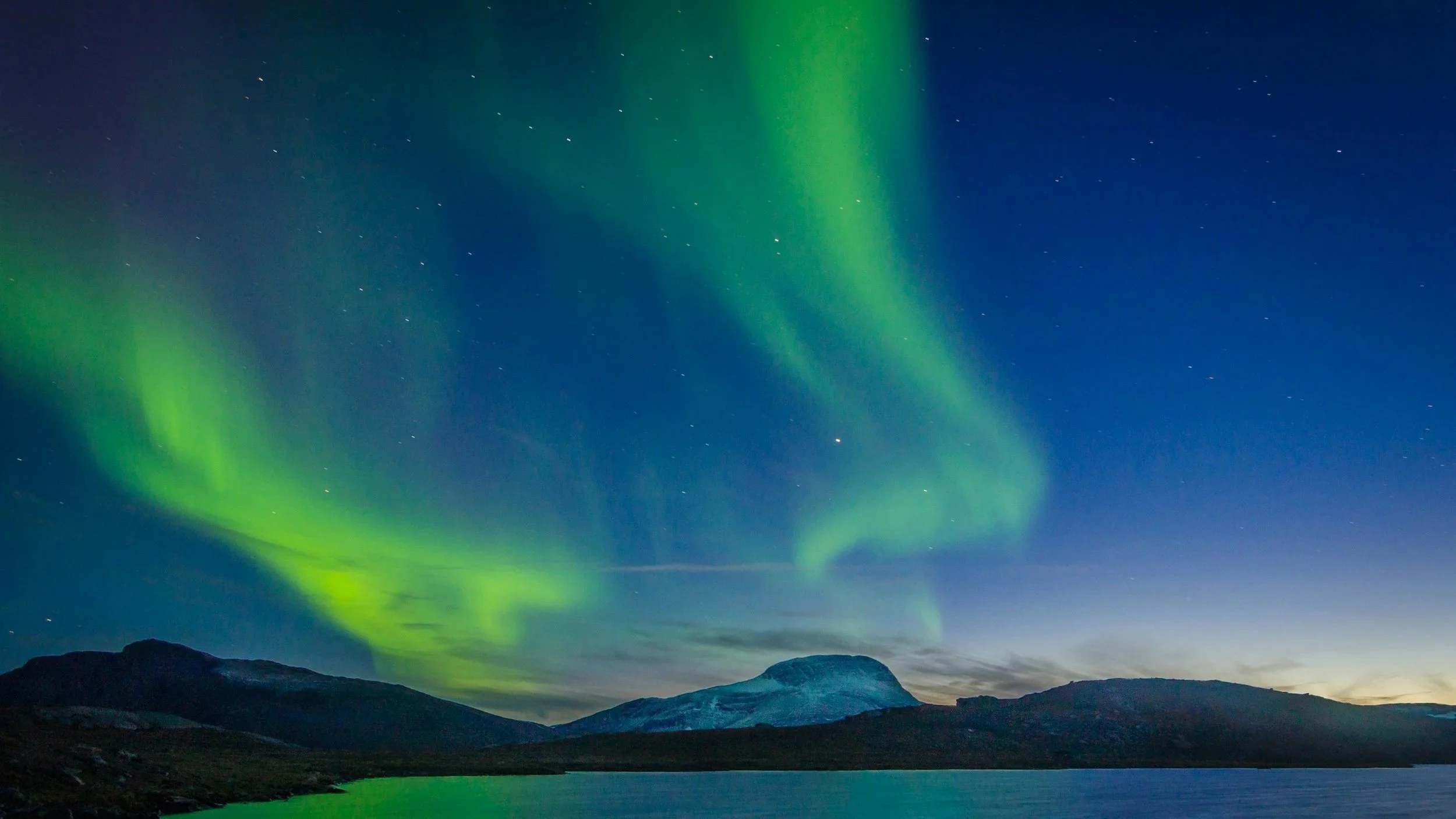 Northern lights (aurora borealis) in the night sky above mountains and a body of water.