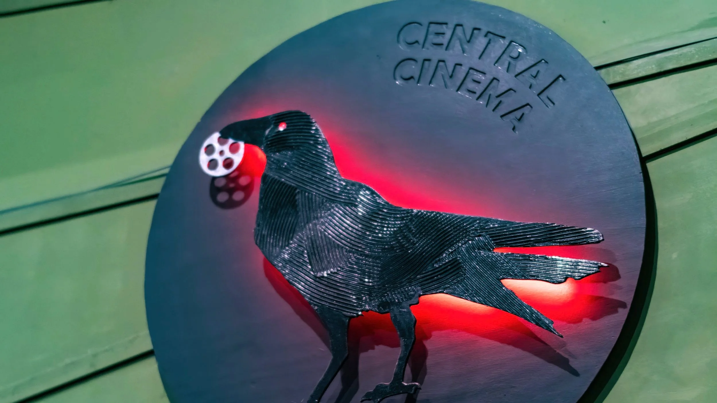 Close-up of a 3D metal sculpture of a crow on a circular surface with engravings, set against a green background.