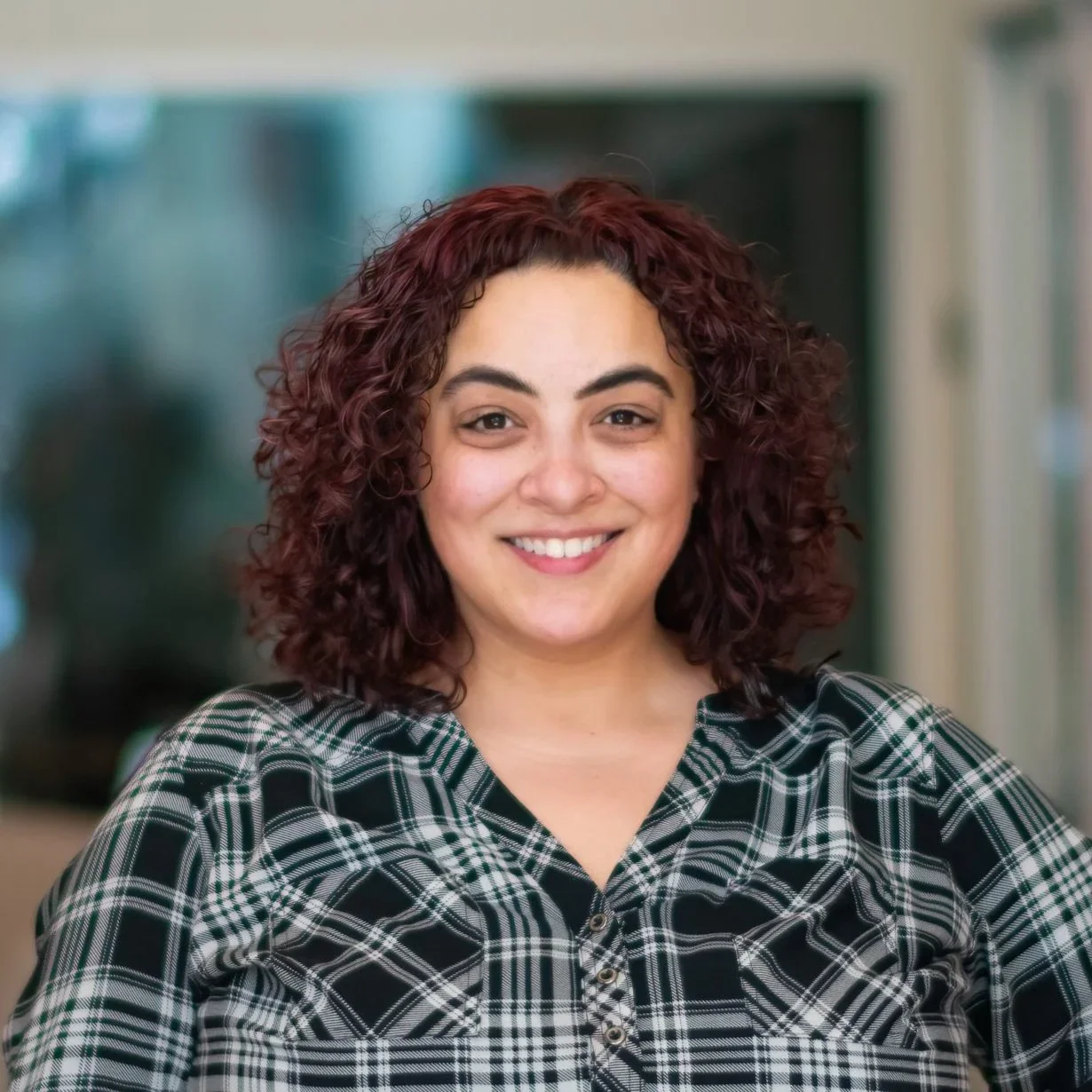 A woman with curly reddish-brown hair smiling, wearing a black and white plaid shirt, in an indoor setting with a blurred background.
