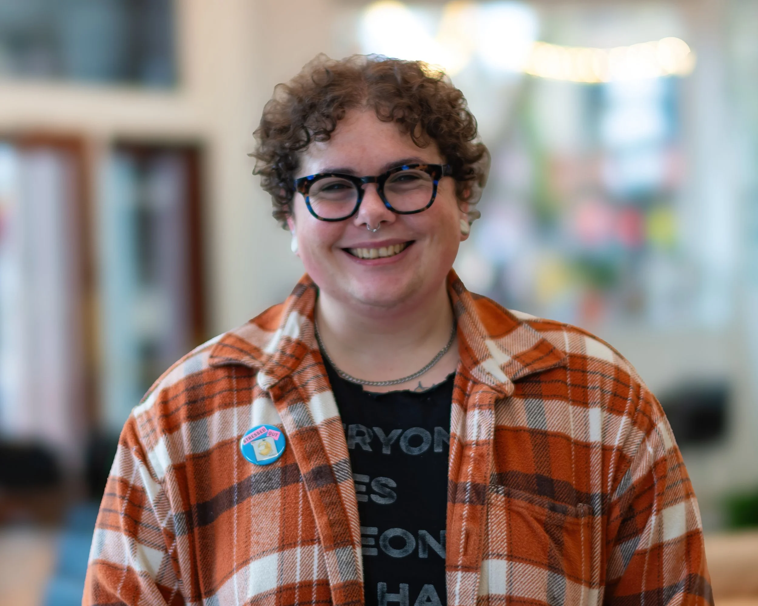 A person with short curly hair, glasses, and a nose ring, smiling indoors. They are wearing a plaid shirt with a sticker on the chest and a black t-shirt underneath, with blurred background.