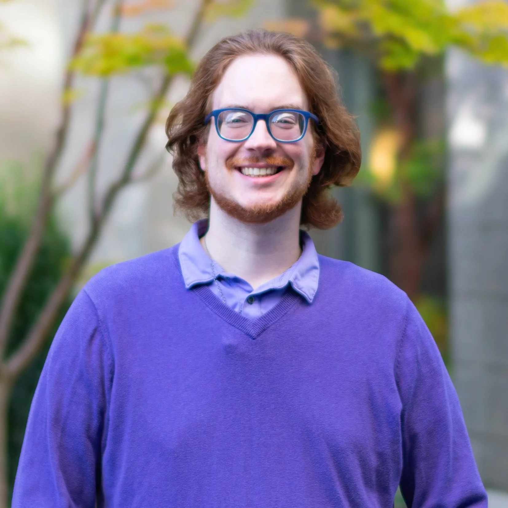 A young man with red hair, glasses, and a beard smiling outdoors, wearing a purple sweater and a light blue collared shirt, with green foliage in the background.