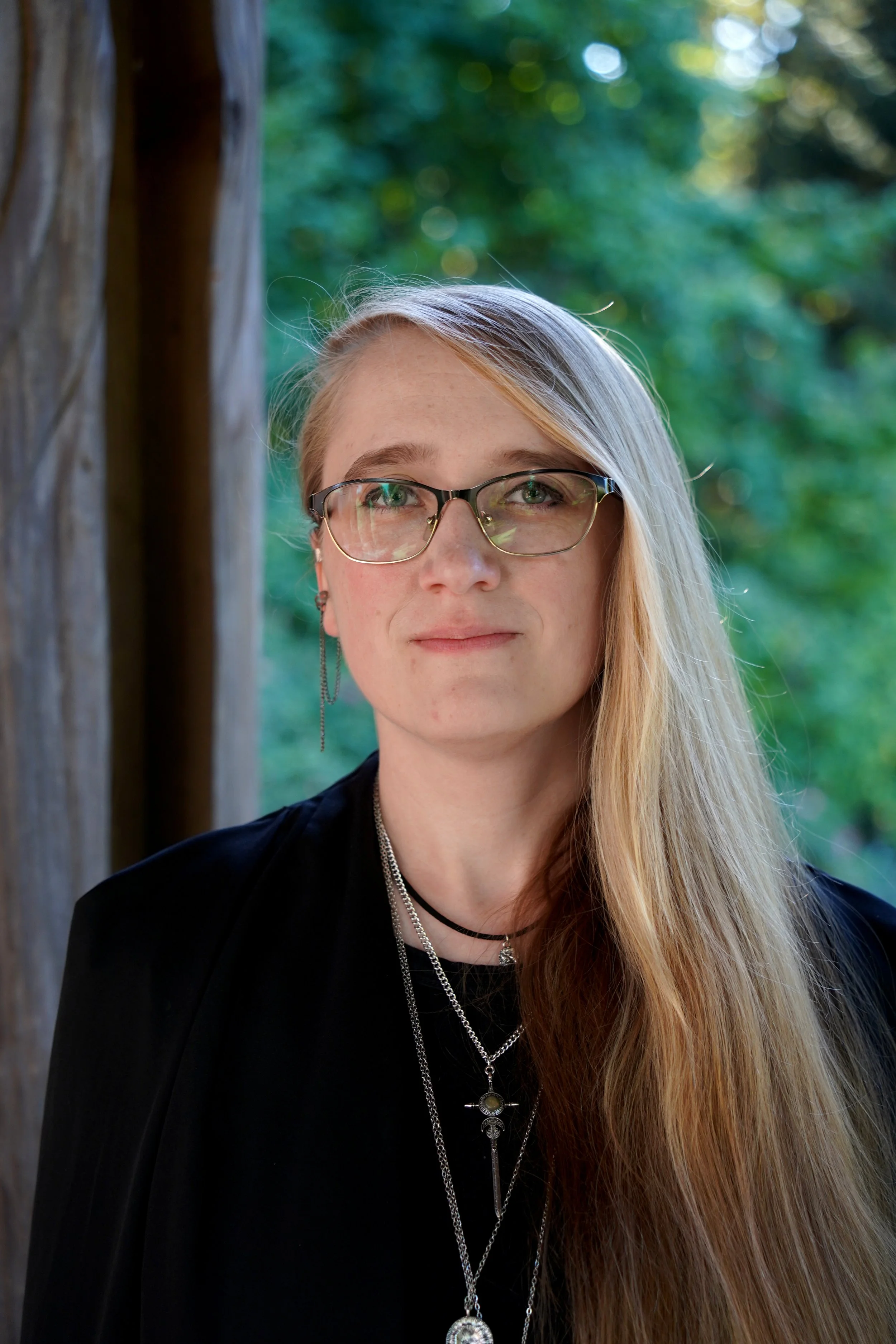 Portrait of a woman with long blonde hair, glasses, layered necklaces, and earrings, standing outdoors near a wooden structure with green trees in the background.
