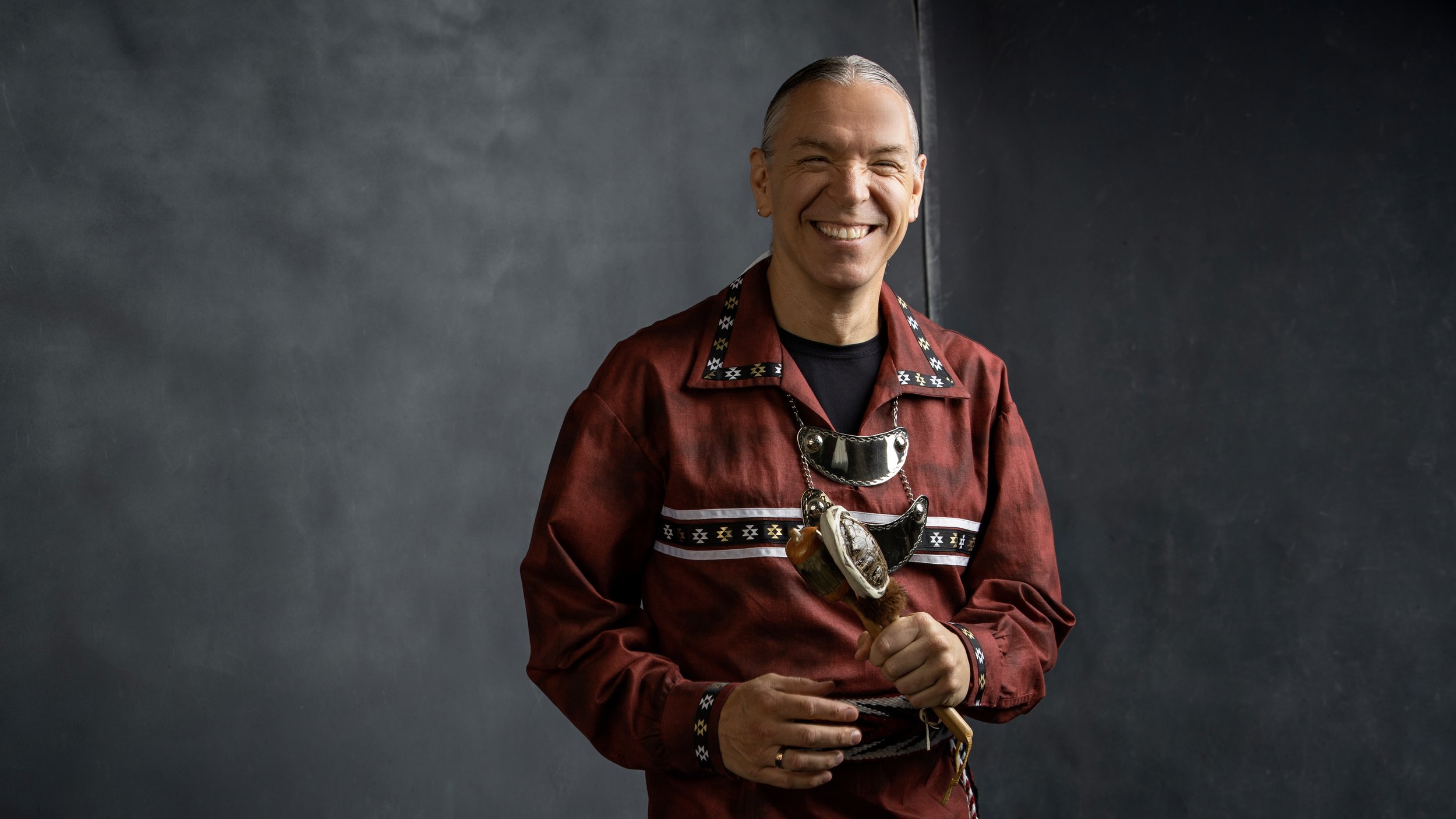 Tate standing and smiling dressed in a rusty red tunic and accessories, holding a wooden instrument.