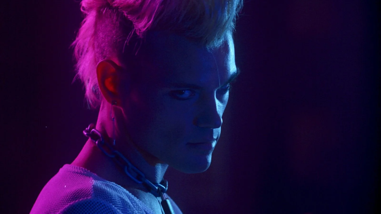 a young masculine person in club lighting wears a chain with a lock around his neck, his expression dark or brooding