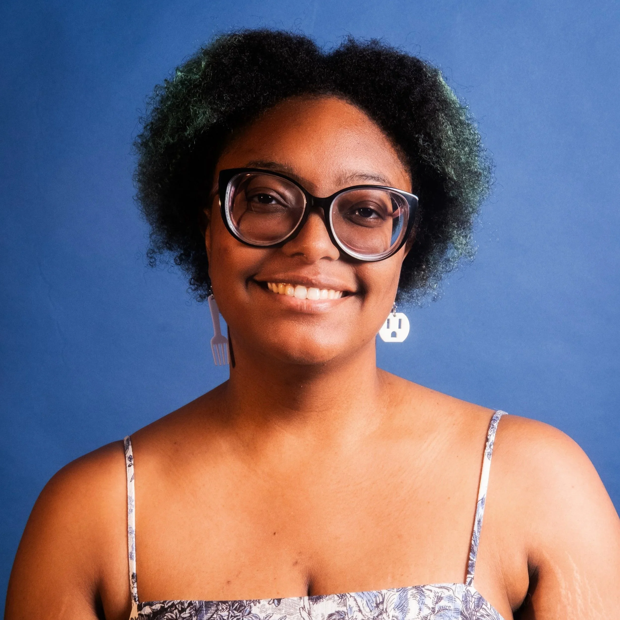 Smiling woman with colorful curly hair, glasses, and floral patterned shirt outside.