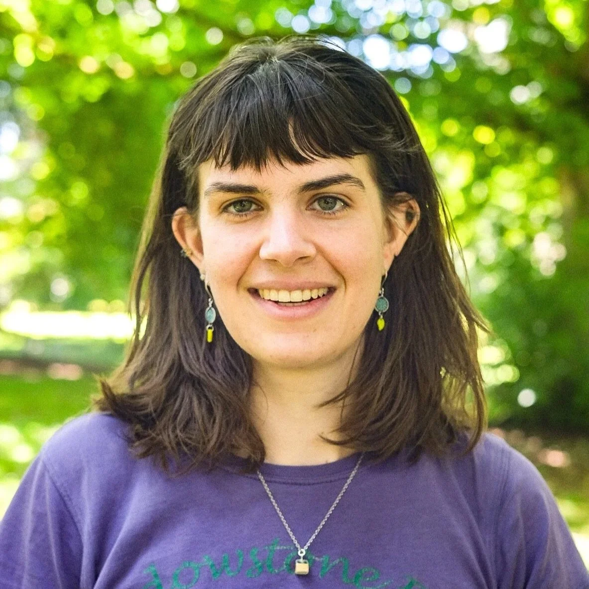 A smiling woman with blue eyes and dark brown hair in an outdoor setting with blurred greenery in the background.