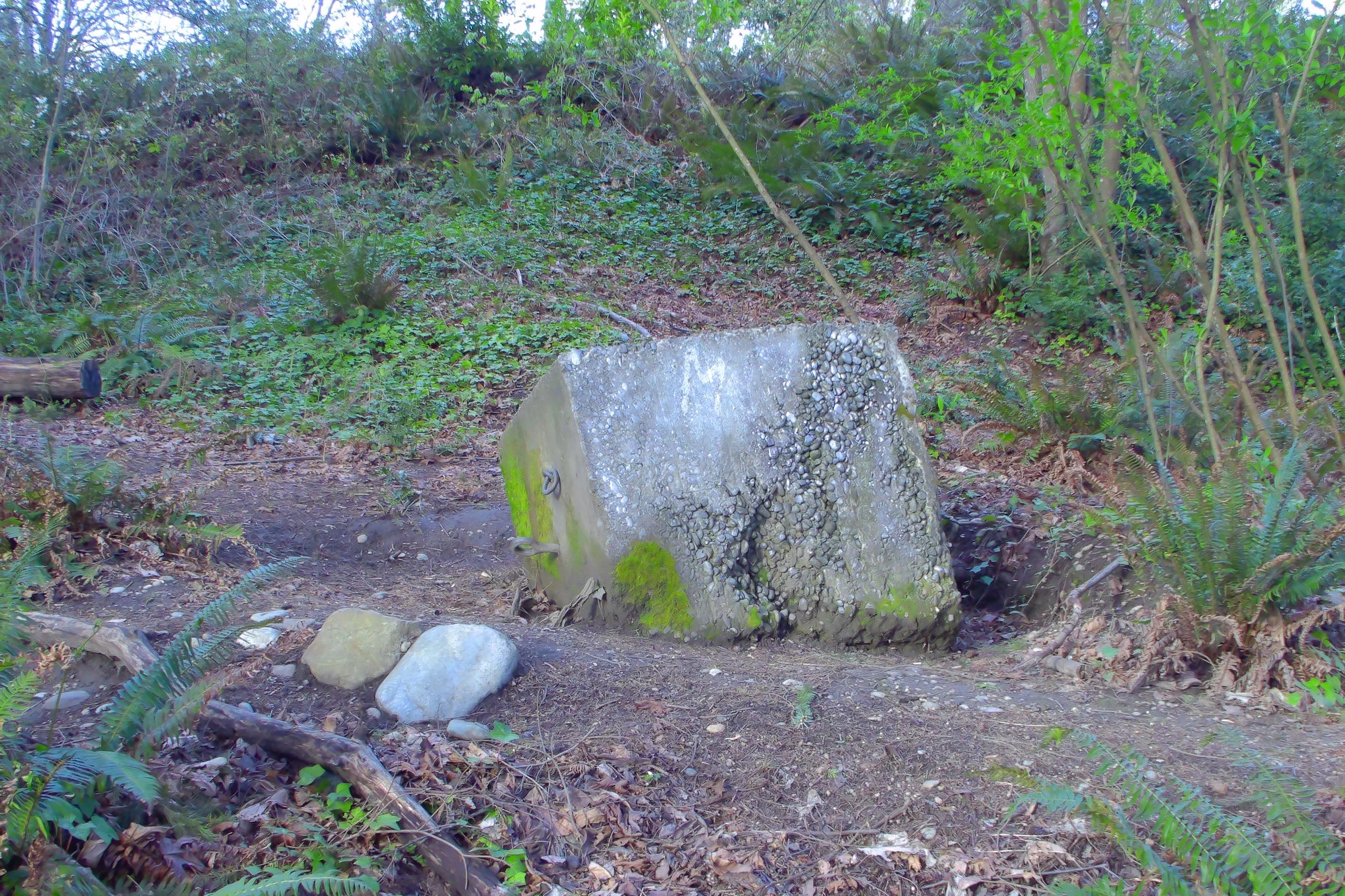 large concrete block with hooks and loops popping out of its side; discarded with moss growing on it at the bottom of a hill.