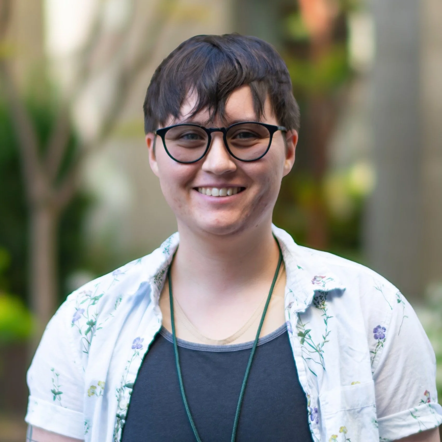 A smiling young person with short dark hair, glasses, a dark shirt, and a light open shirt with a floral pattern, outdoors with blurred trees and greenery in the background.