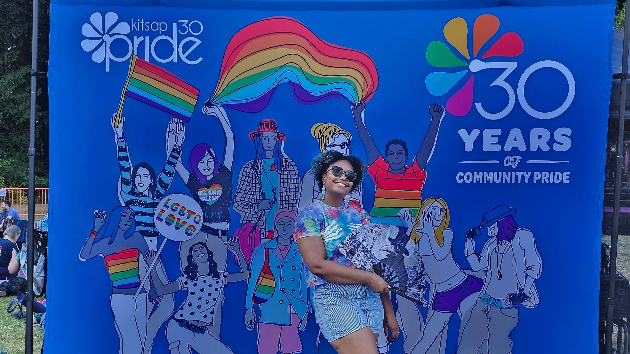 Raegan in front of the Kitsap Pride blue backdrop illustrated with rainbows and people