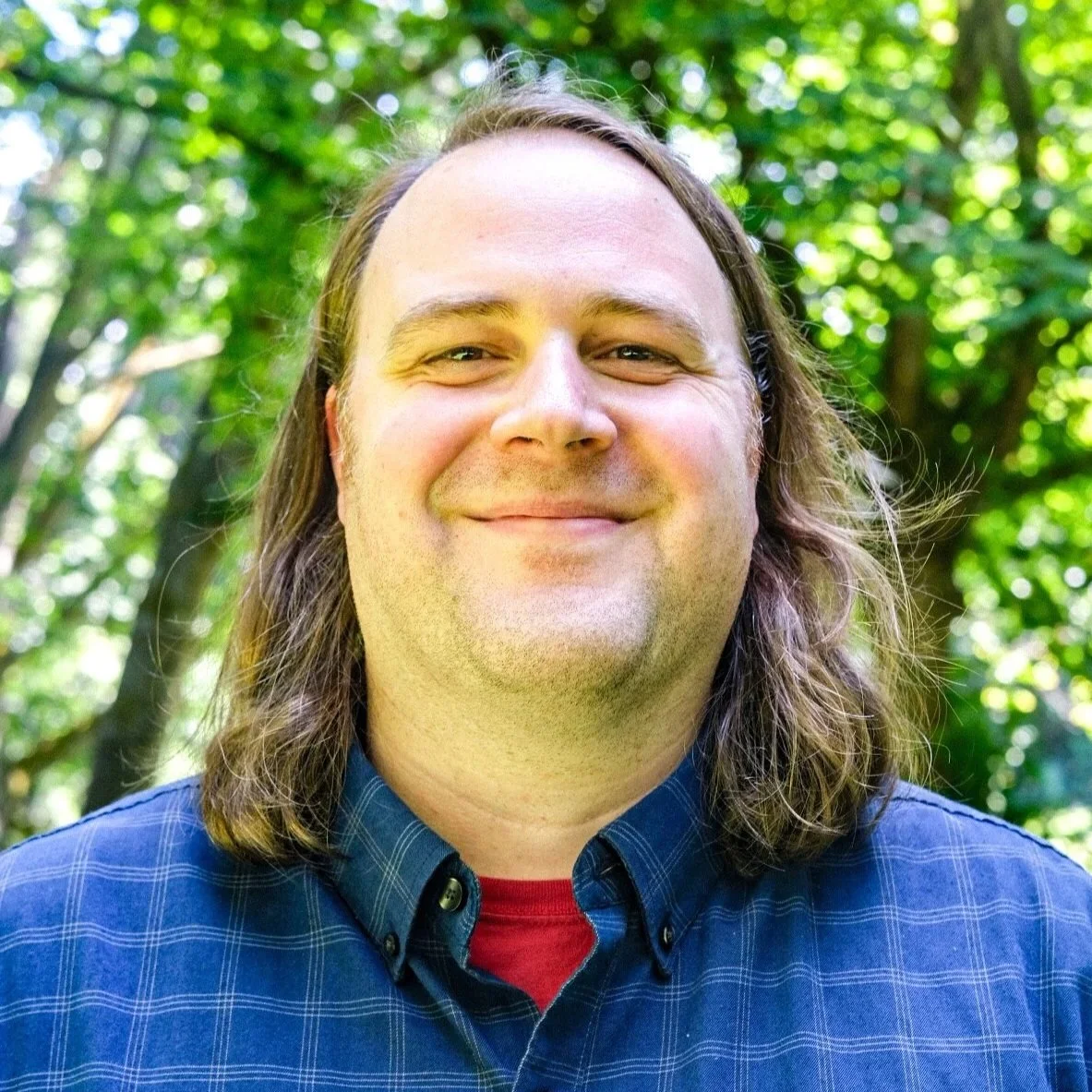 A smiling man with light brown hair, a beard, and mustache, wearing a green plaid shirt, standing indoors against a blurred background.