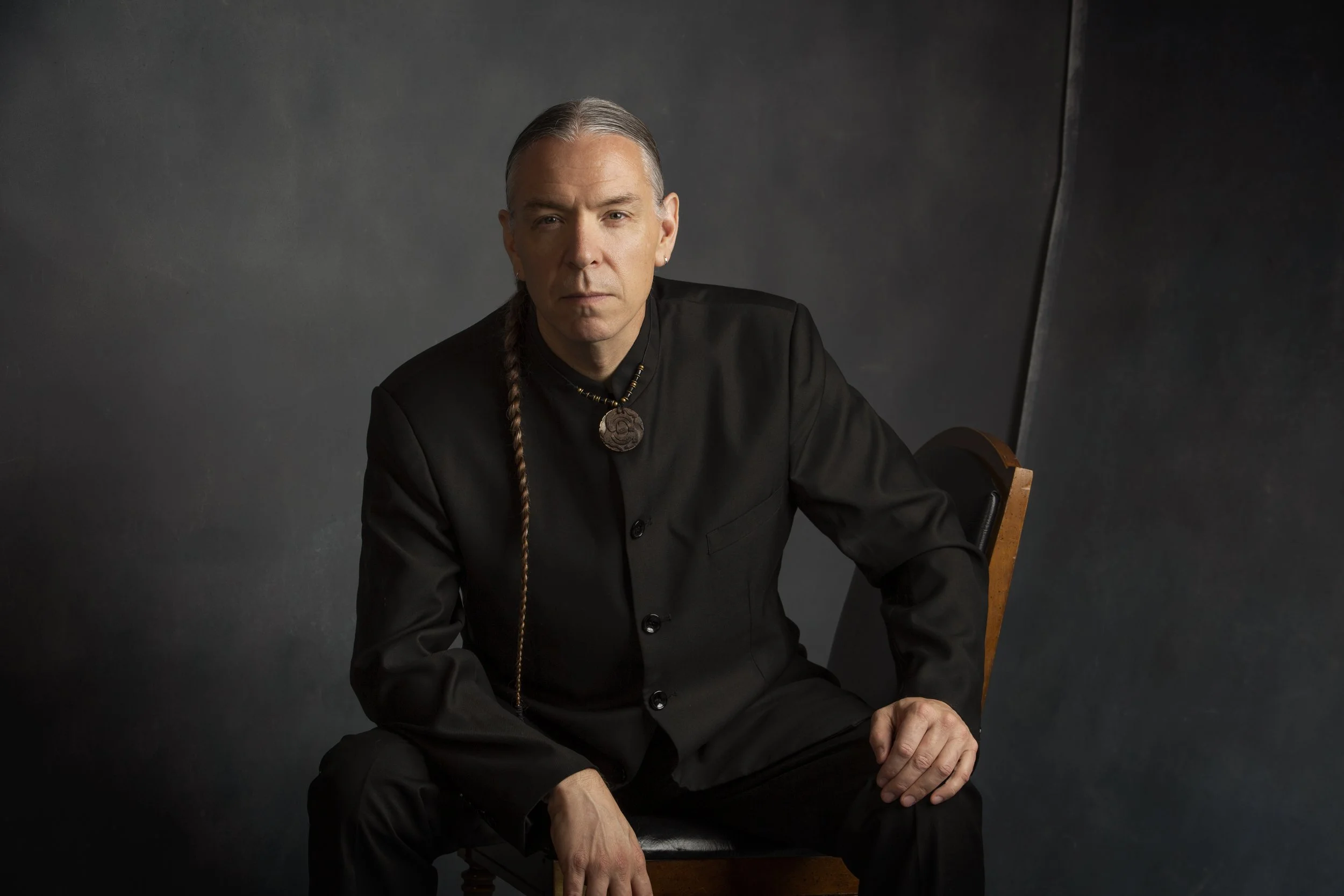Jerod Impichchaachaaha’ Tate seated in a wooden chair, dressed in black