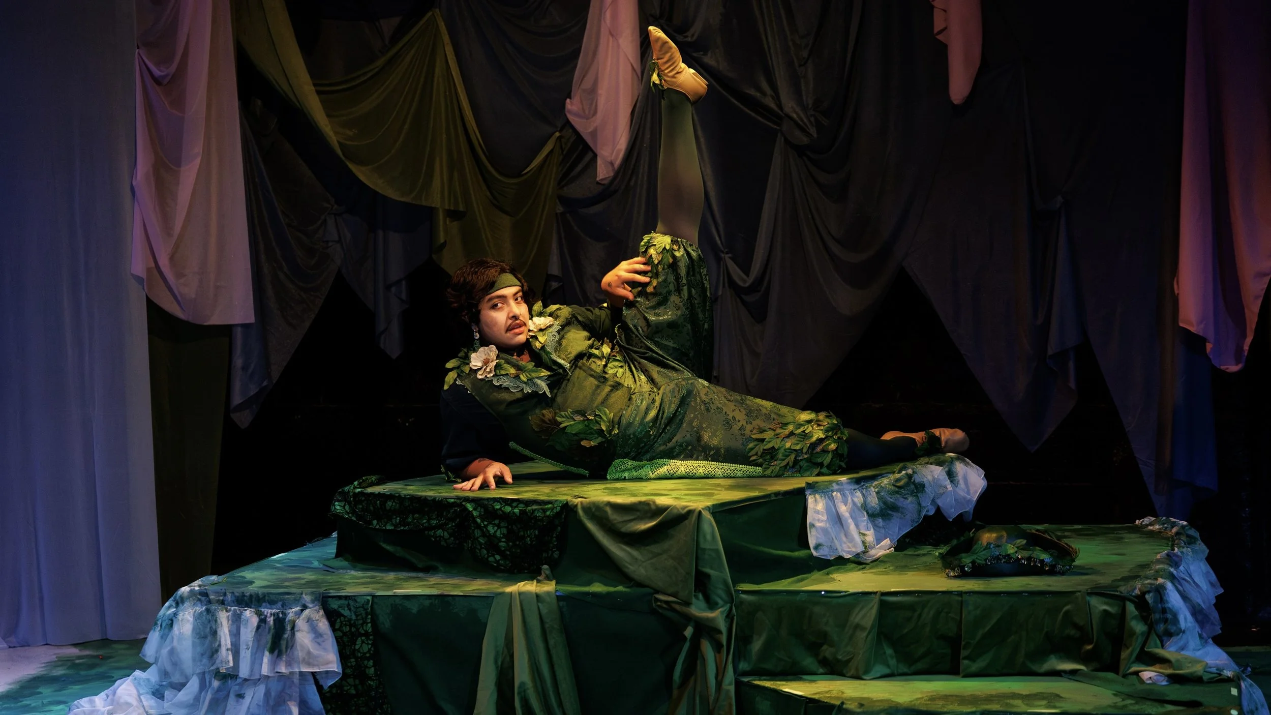 Character Shelmerdine in Orlando, lying atop 3 tiers on their side with left leg raised to the sky, dressed in green with floral accents