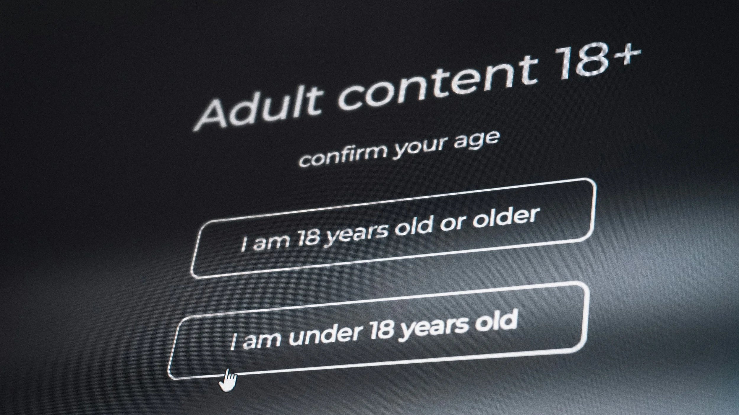grayscale photo of a computer monitor with prompt for "adult content 18+" and "confirm your age" with pointer on button for Under 18