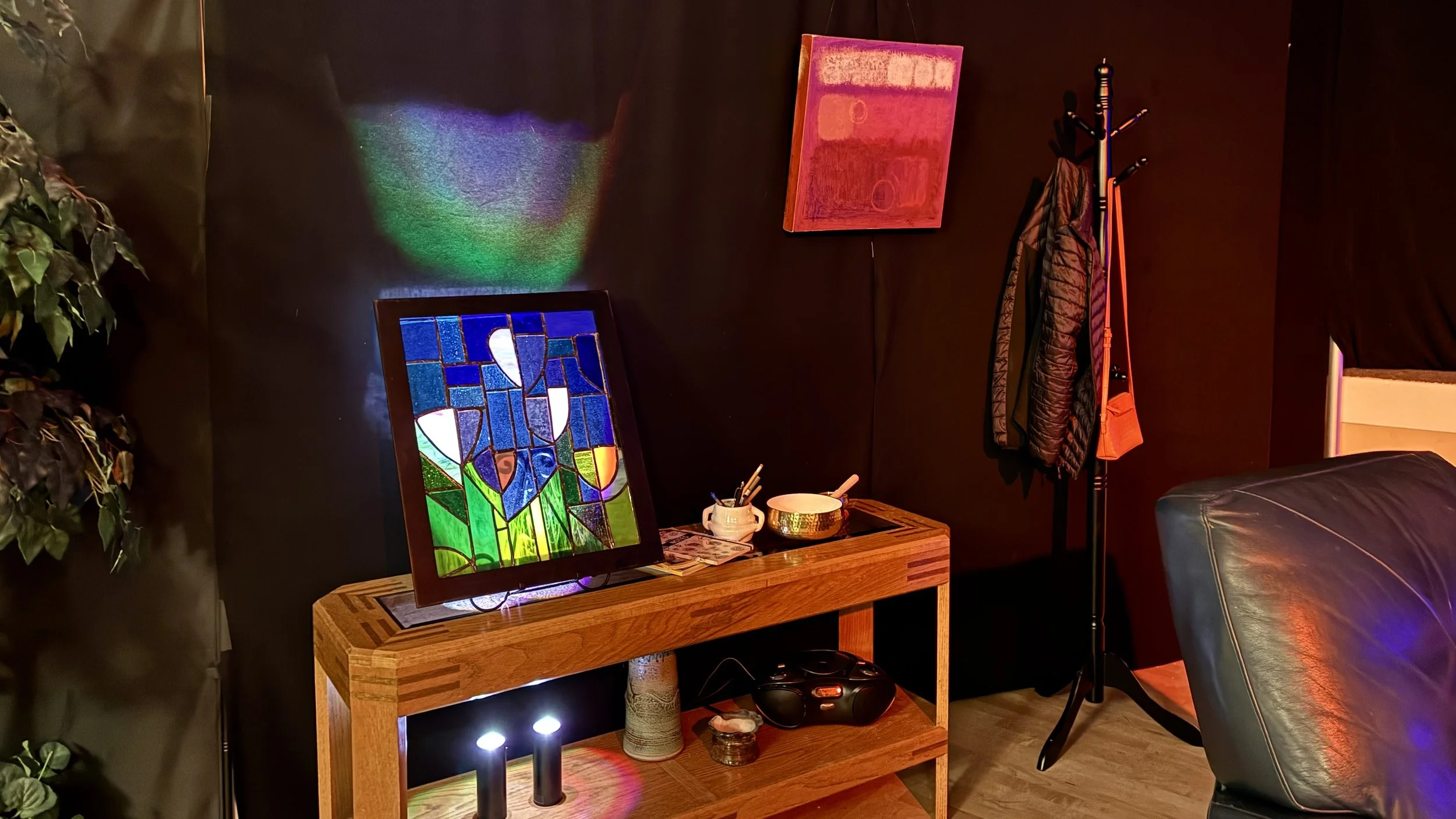 set designed as a portion of a living room, with a coat hanging on a rack, a shelf with knickknacks, framed stained glass; brown walls with a hung painting; dark leather chair