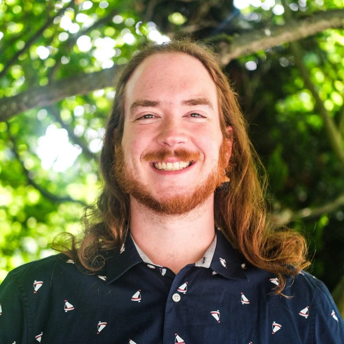 A young man with red hair, glasses, and a beard smiling outdoors, wearing a purple sweater and a light blue collared shirt, with green foliage in the background.