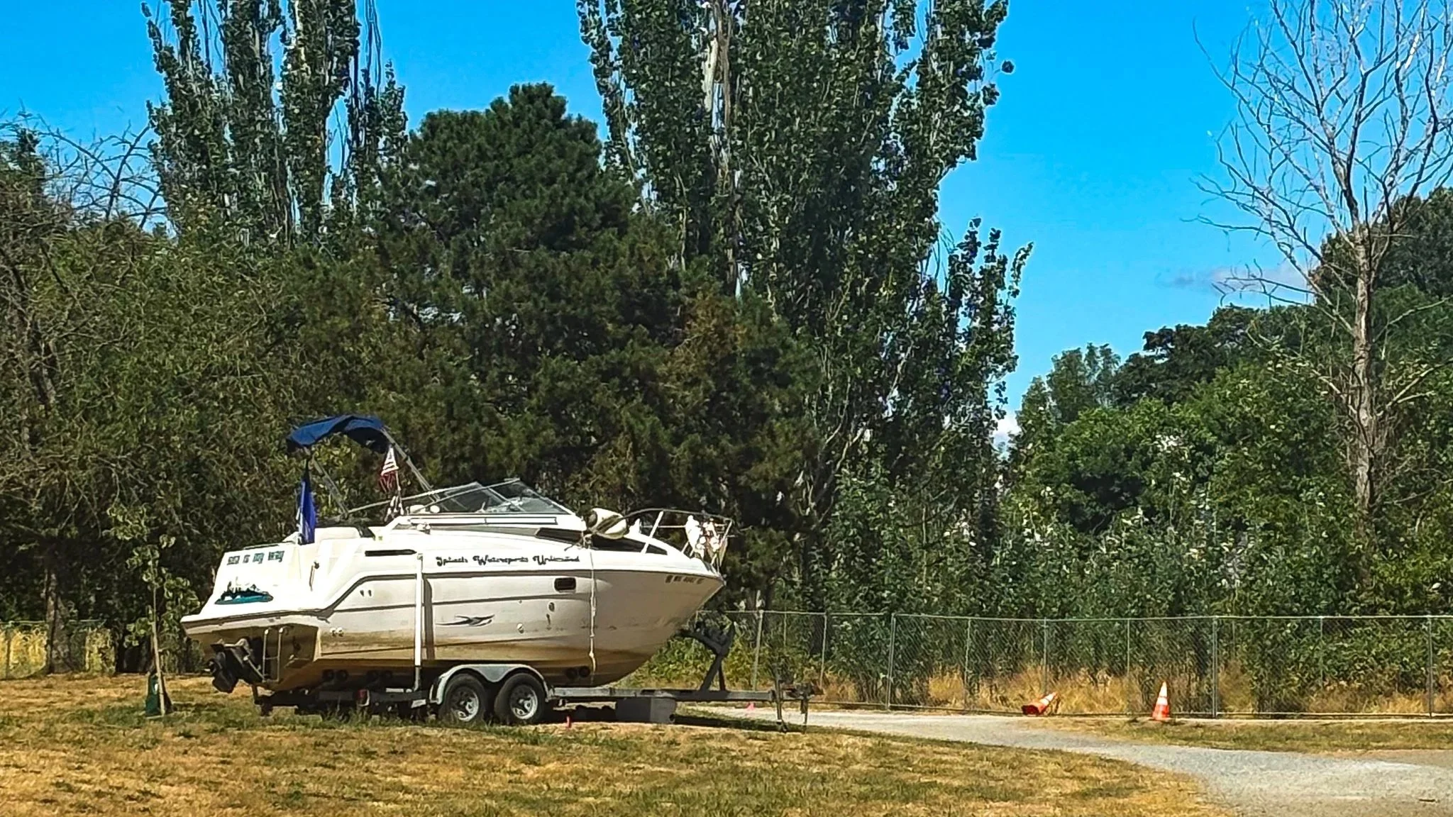 white boat on wheels parked at a trail across from a fence lining woods