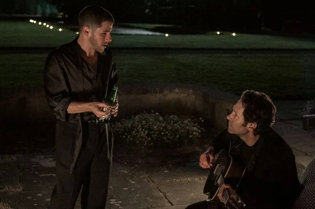 Still from Power Ballad; one man stands while chatting with the seated man played by Paul Rudd holding a guitar