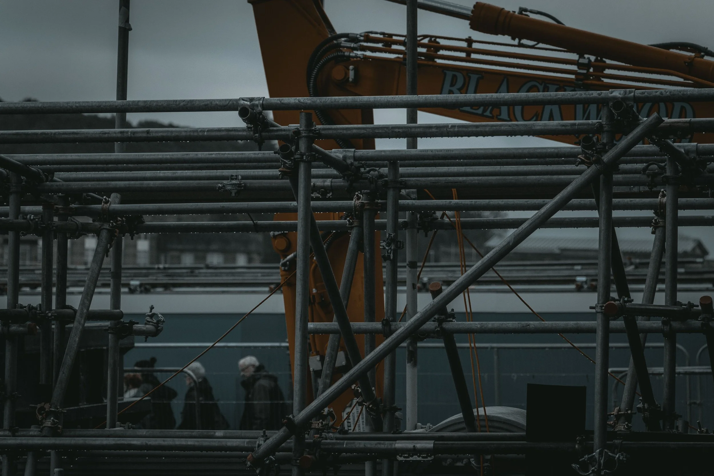 Metal pipes form a structure. People walk far behind it and closer behind the structure is the orange arm of a construction vehicle