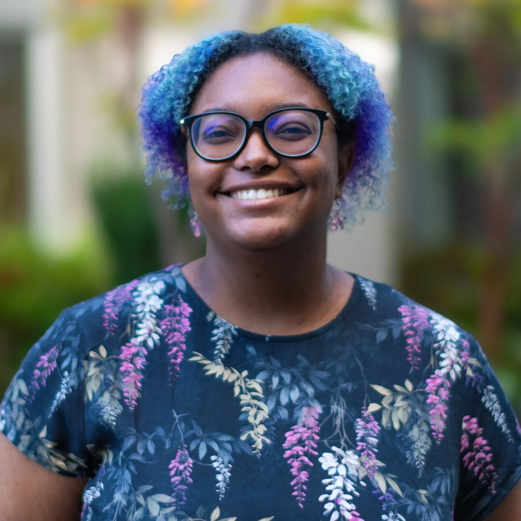 Smiling woman with colorful curly hair, glasses, and floral patterned shirt outside.