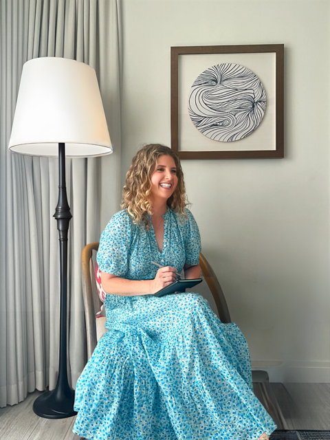 Felicia Andrews, LCSW, smiling while seated in a blue floral dress, with a floor lamp, curtains, and framed artwork in the background.