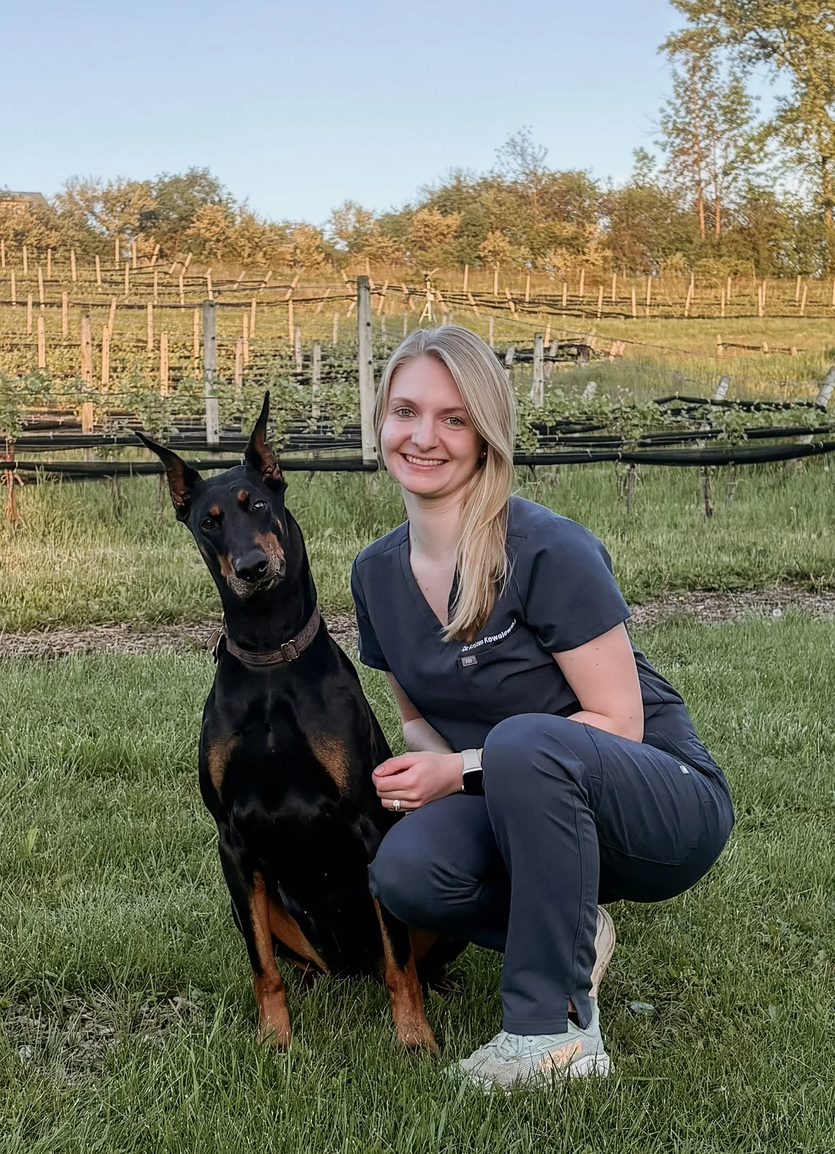 Dr. Kowalewski in navy blue scrubs crouched next to her doberman dog in front of a field