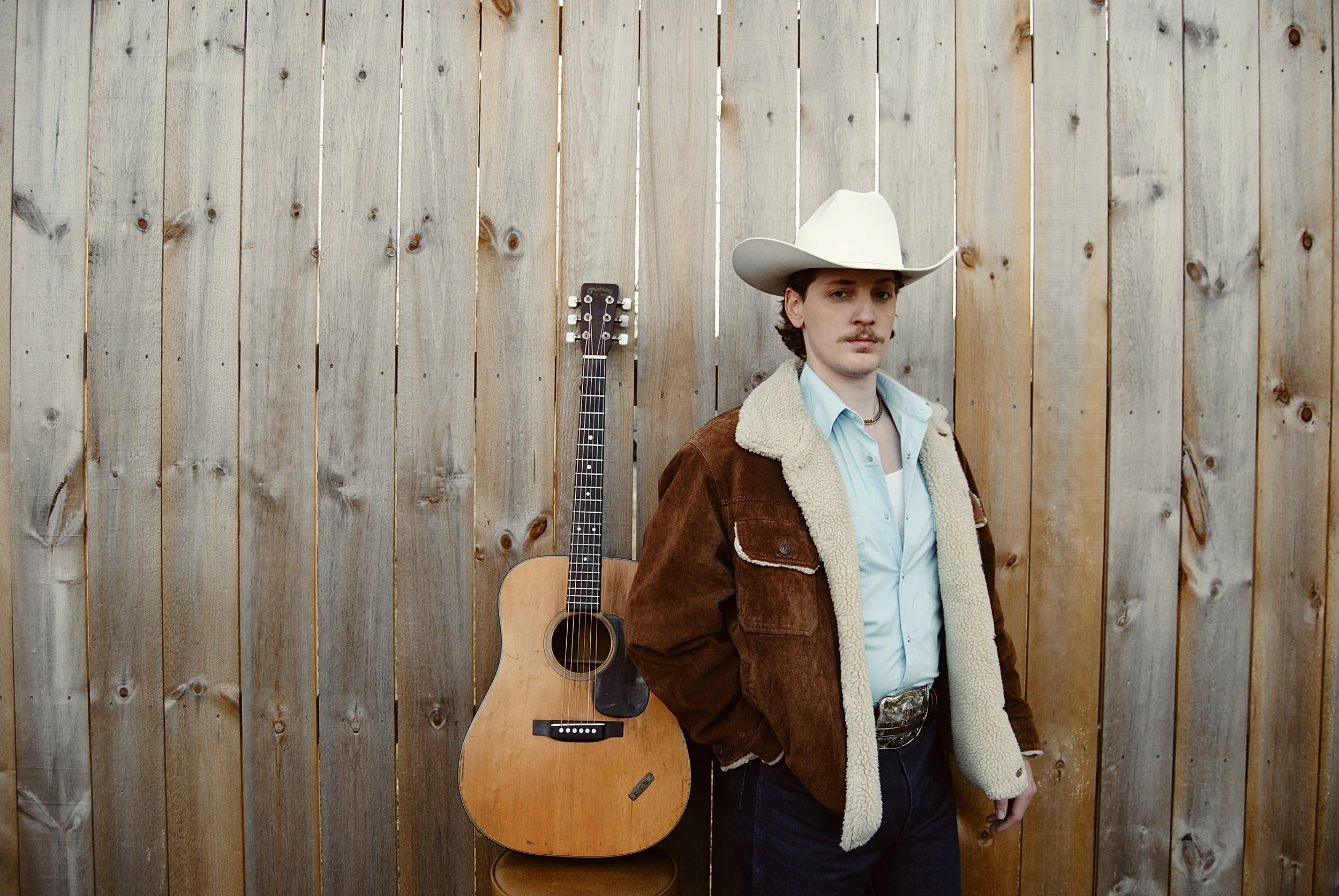 A man wearing a cowboy hat and Western-style jacket standing next to an acoustic guitar against a wooden fence.