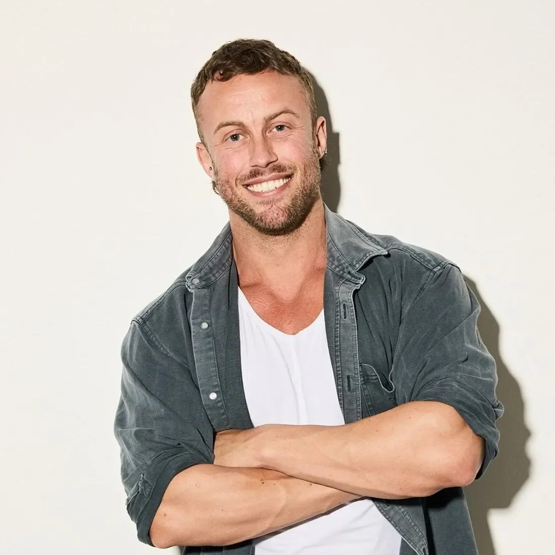Steve Spencer smiling with arms crossed, wearing gray denim shirt over white v-neck tee. He has short brown hair, light facial hair, and ear piercings.