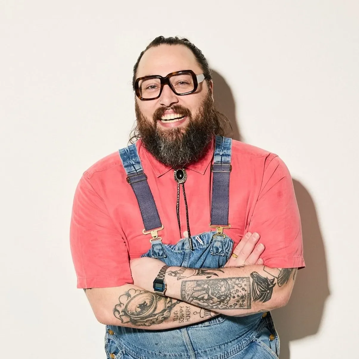 Chad D'Elboux Barnier smiling with arms crossed, wearing large black-framed glasses, coral pink shirt, denim overalls, bolo tie, and displaying tattoos on both forearms.