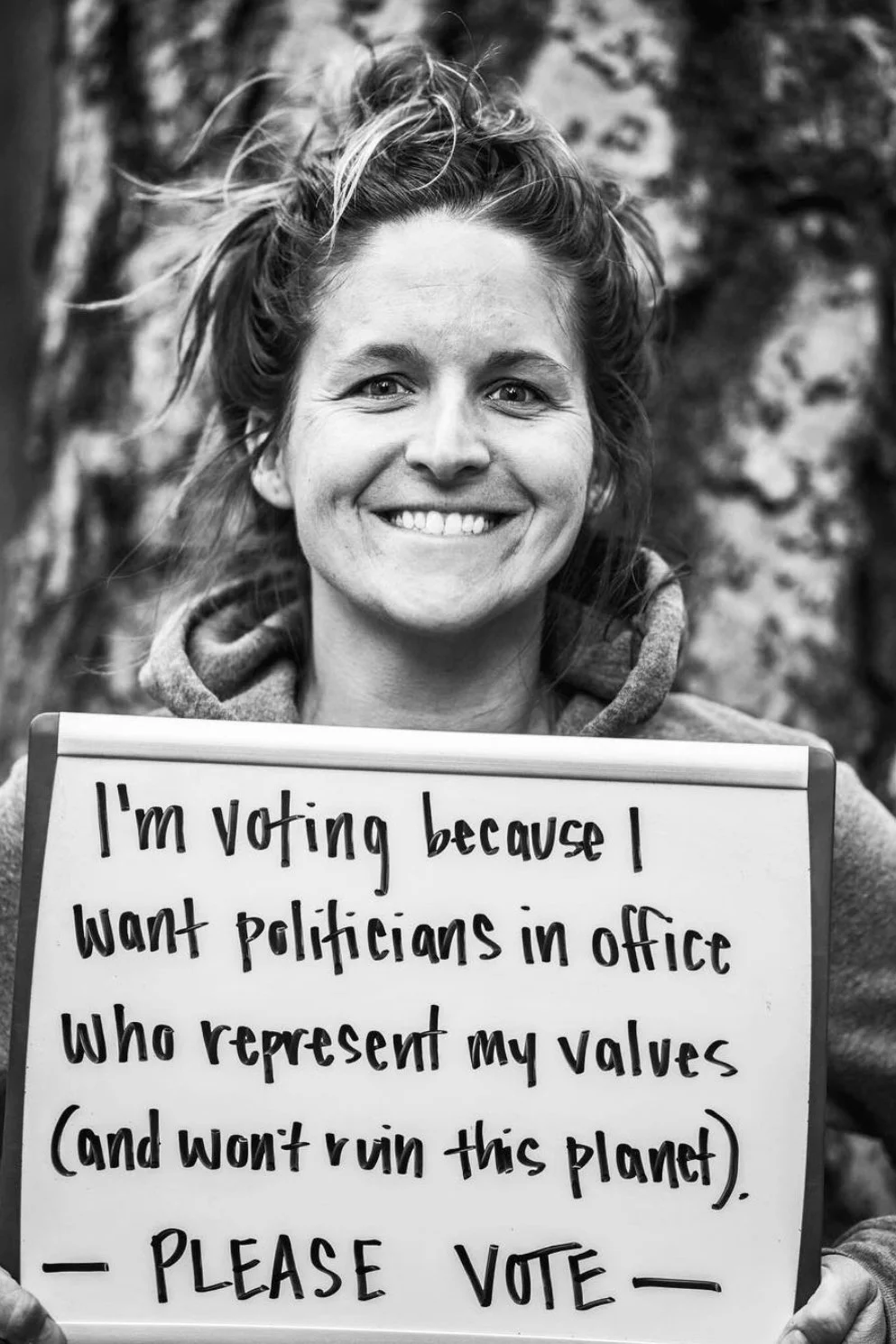 A woman with tousled hair smiling and holding a sign that says "I'm voting because I want politicians in office who represent my values (and won’t ruin this planet). - PLEASE VOTE." The background appears to be outdoors with blurred trees.