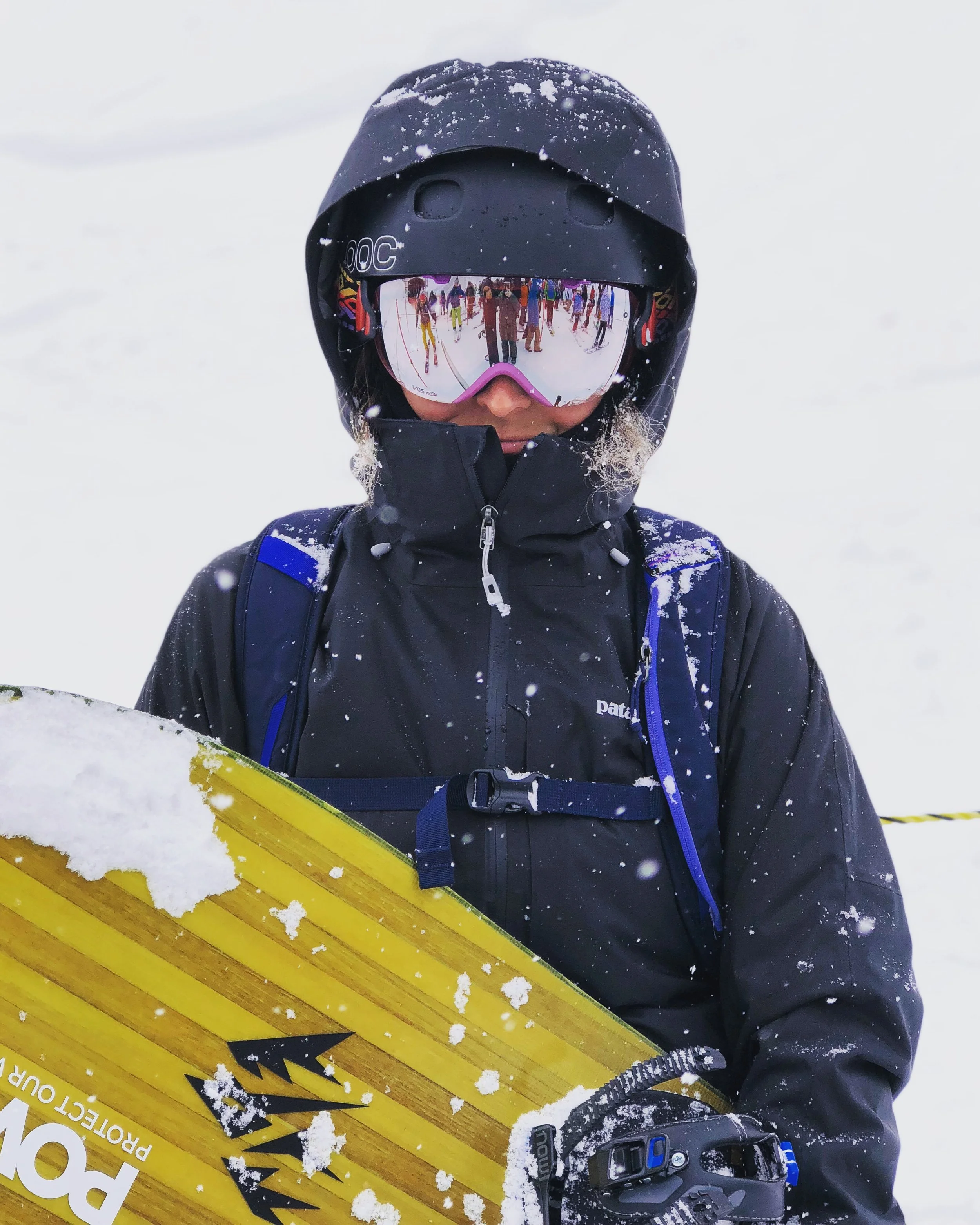 Person dressed in winter gear and ski goggles holding a yellow snowboard in a snowy landscape with a group of skiers reflected in the goggles.