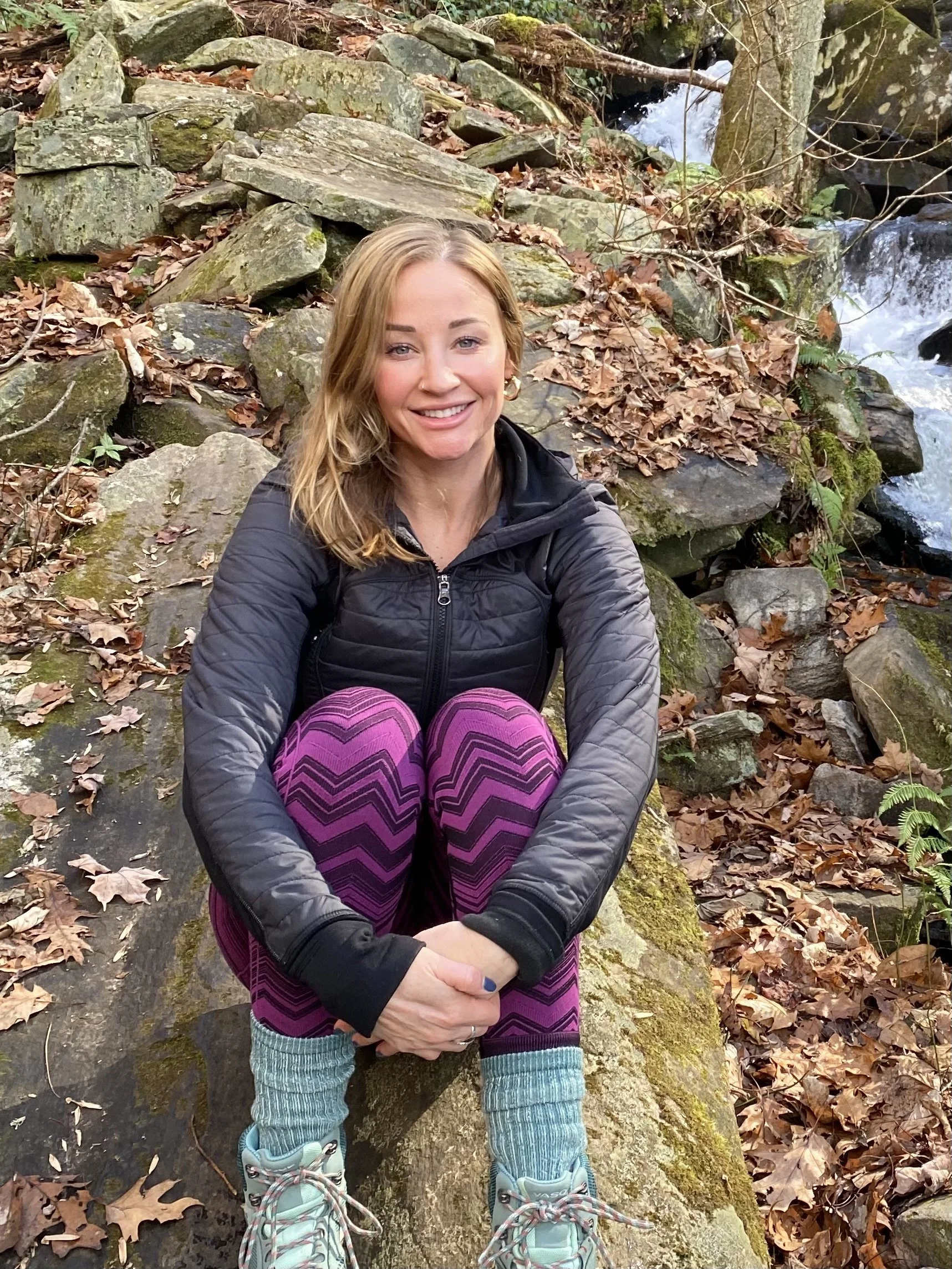 A woman sitting on a moss-covered rock in a forest near a small stream, surrounded by fallen leaves, smiling at the camera.