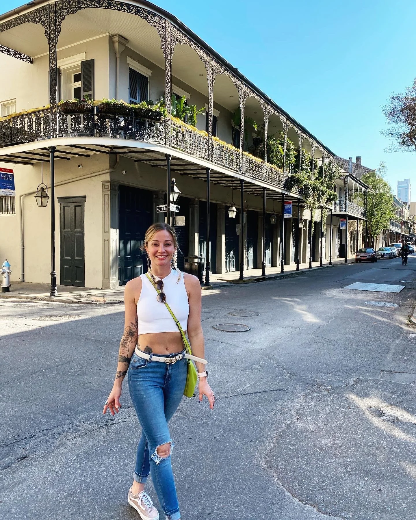 A young woman with blonde hair, wearing a white crop top, ripped jeans, and sneakers, walking on a city street with historic buildings in the background under a clear blue sky.