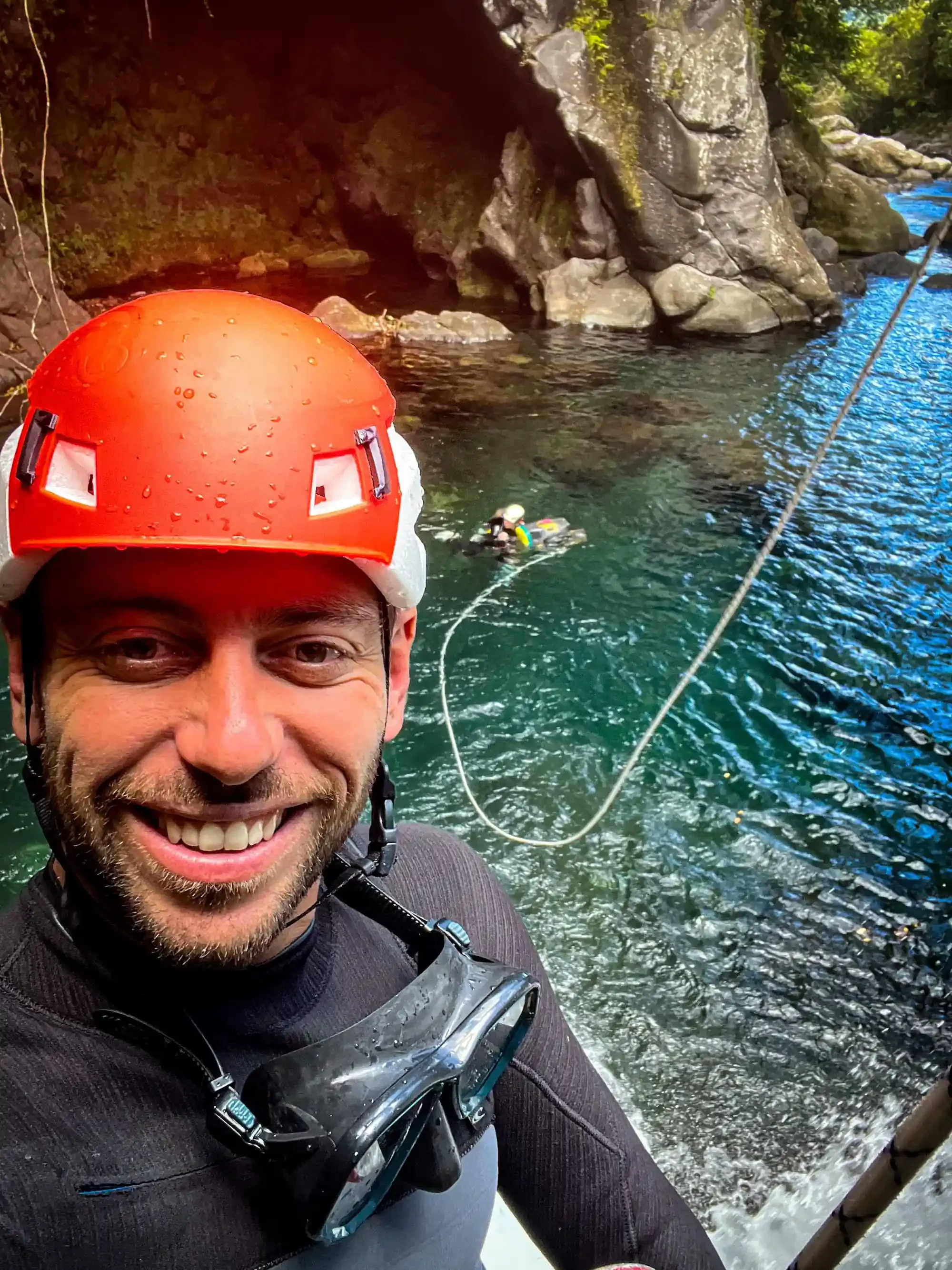 Homme souriant portant un casque rouge, des lunettes de plongée, et une combinaison de sport, en pleine activité de canyoning ou d'escalade dans une rivière entourée de rochers et de végétation.