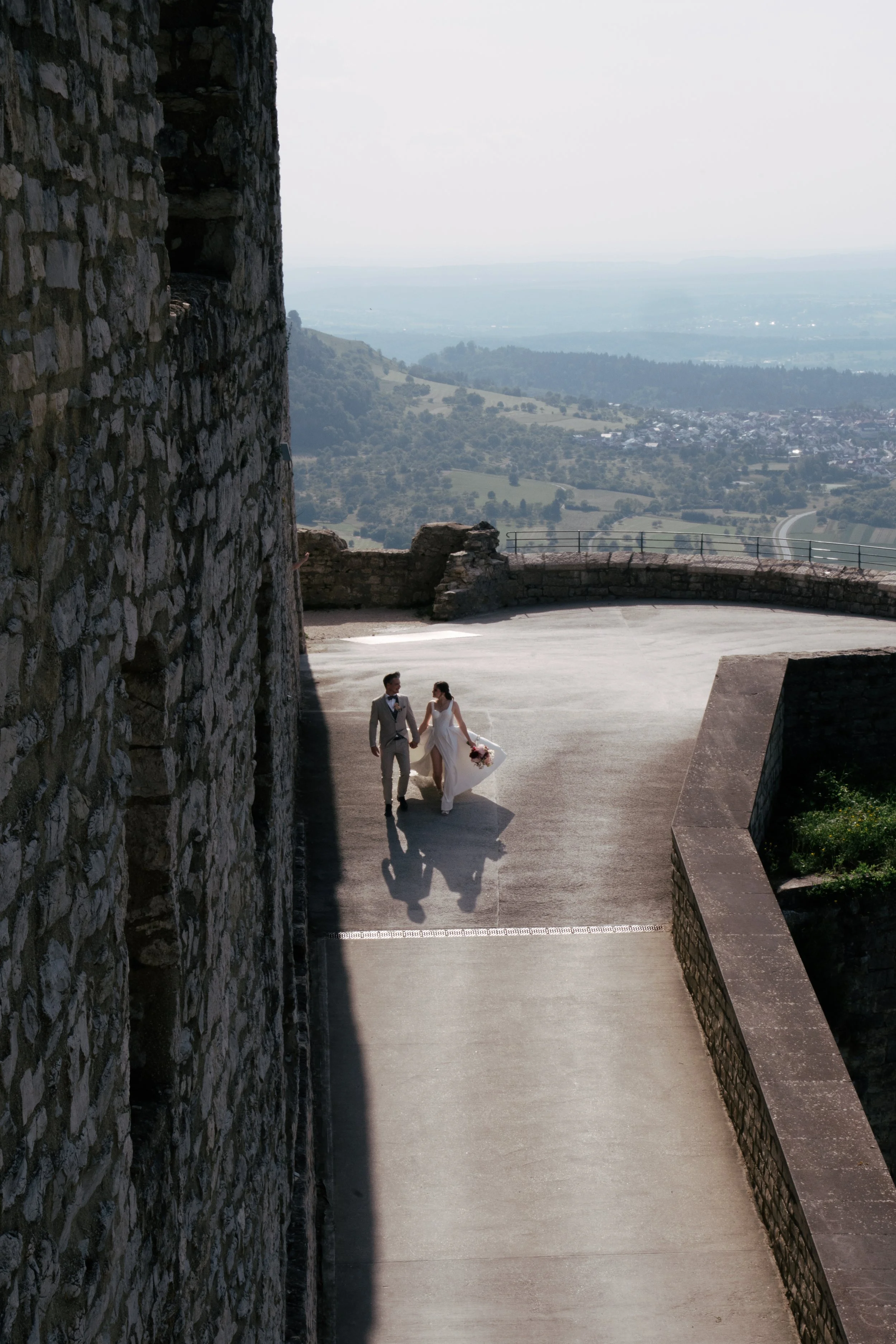Ein Brautpaar läuft Hand in Hand auf einer Terrasse vor einer alten Steinmauer mit Panorama-Ausblick auf das ländliche Gebiet mit Hügeln und Feldern in der Ferne.