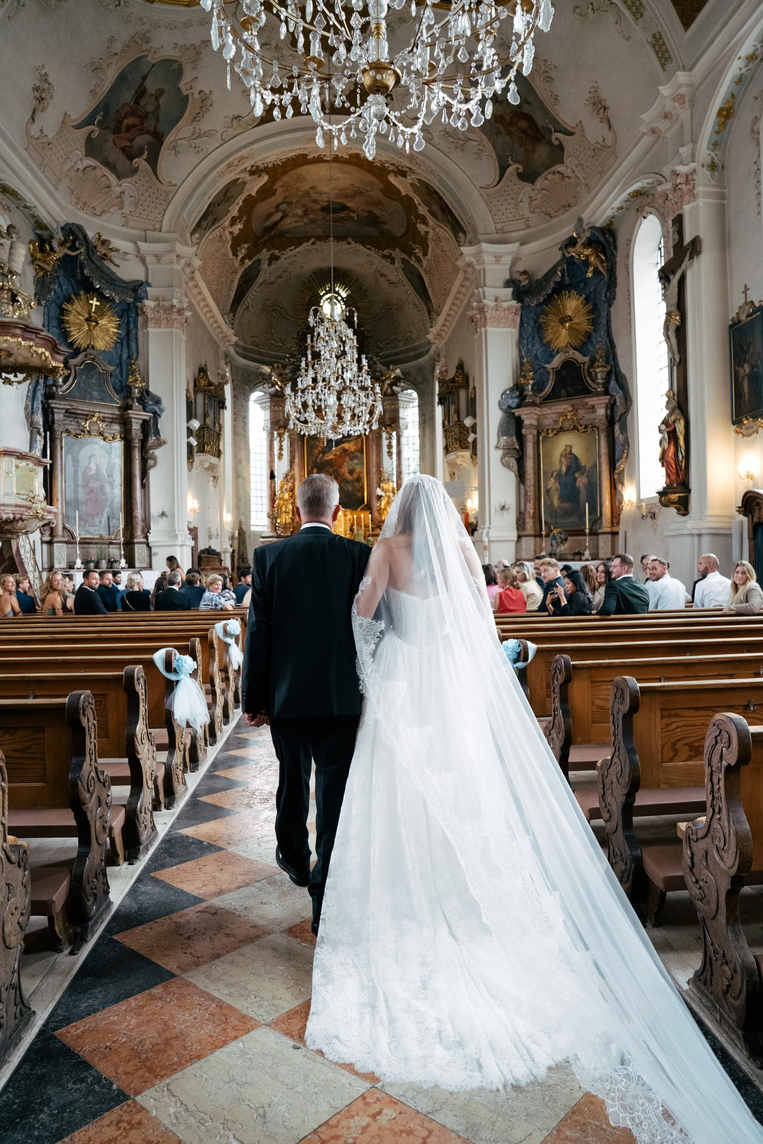 Braut und Bräutigam gehen in einer prachtvollen Kirche zum Altar, umgeben von vielen Gästen, mit Kronleuchtern, goldenen Verzierungen und religiösen Bildern.