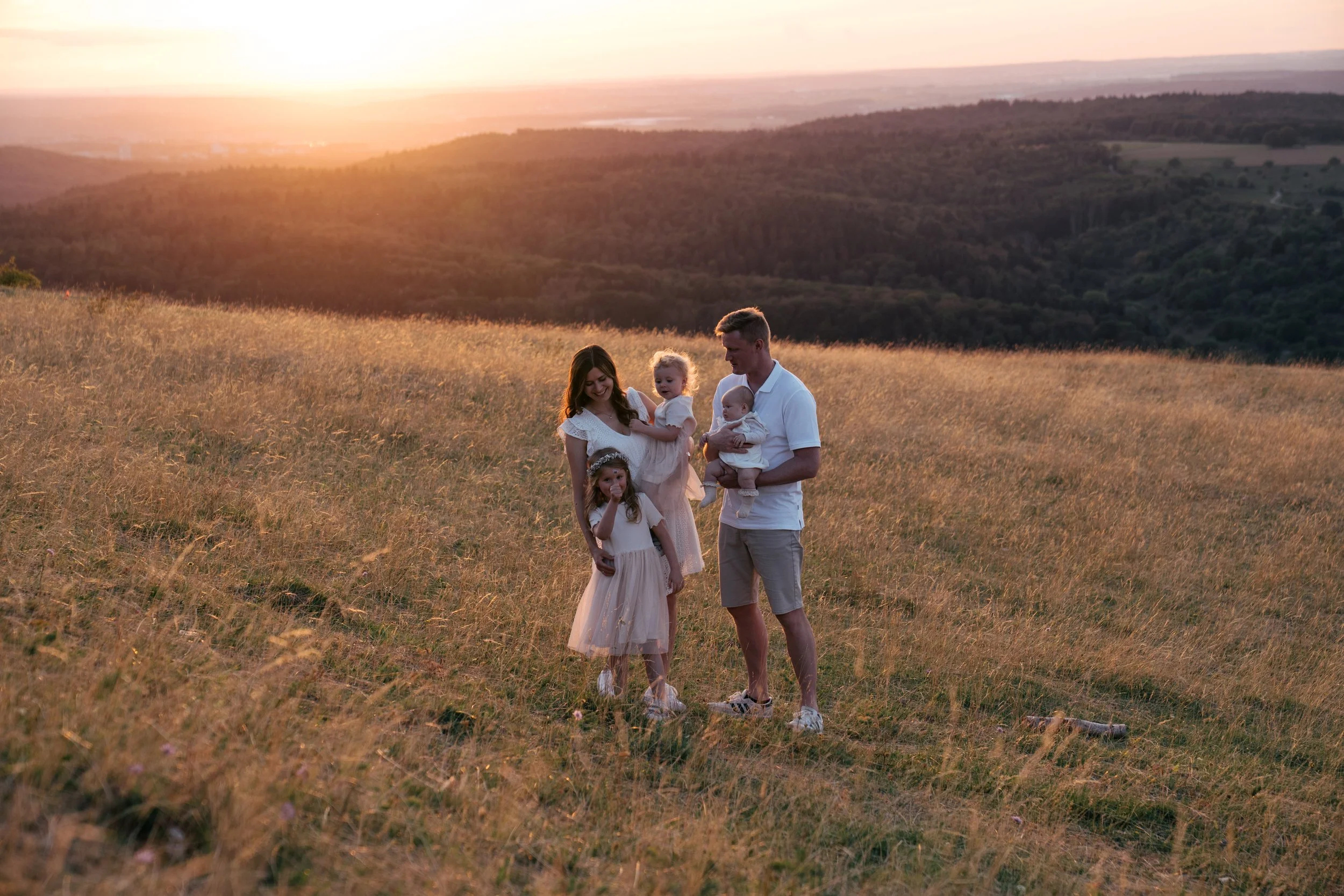 Familie, Mutter, Vater und drei Kinder, wandern in einer grünen, hügeligen Landschaft bei Sonnenuntergang.
