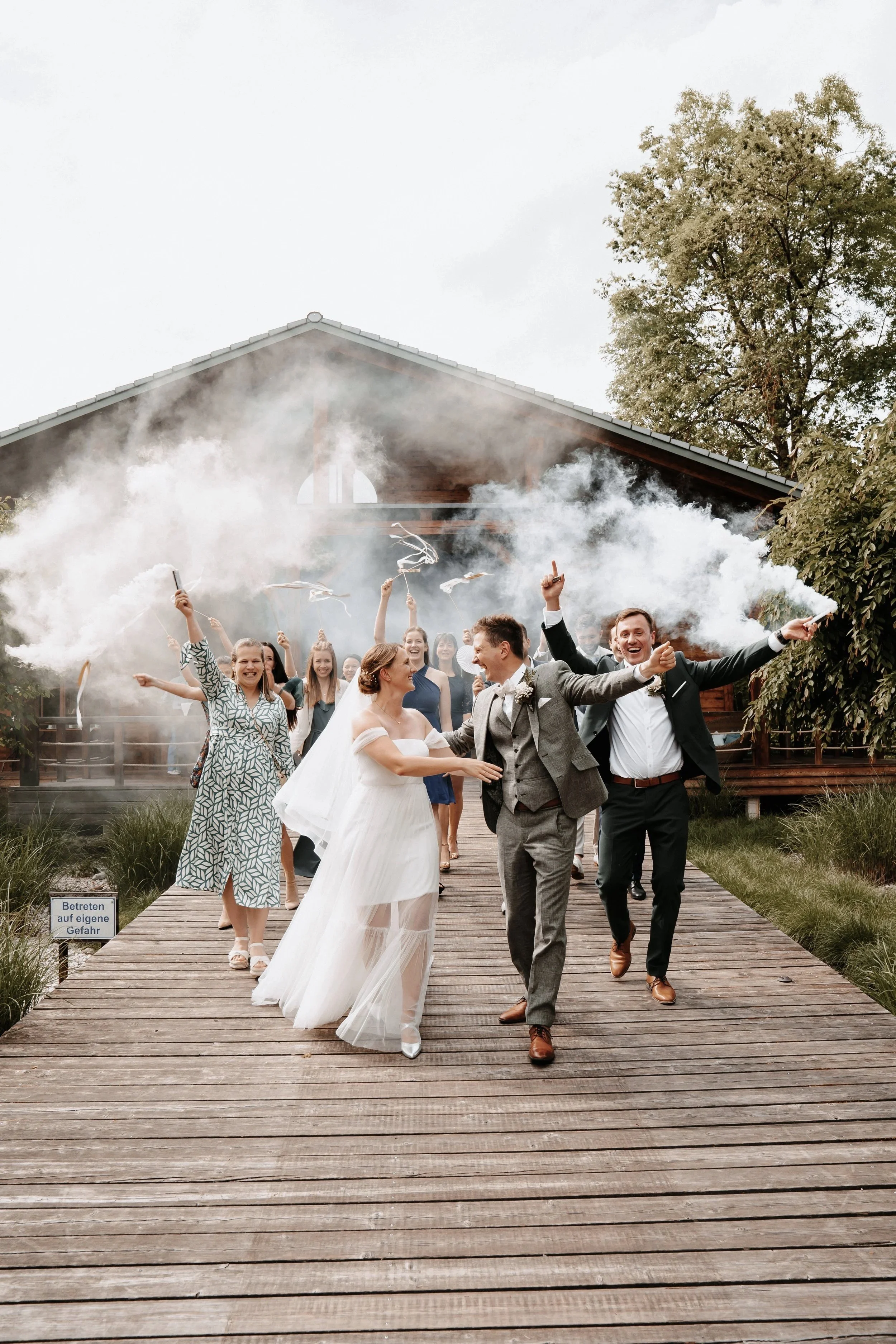 Hochzeitspaar auf einem Holzweg, gefolgt von Feiern mit Rauchfackeln. Gebäude im Hintergrund, Bäume und Gras umgeben die Szene. "Betreten auf eigene Gefahr"-Schild am Rand.