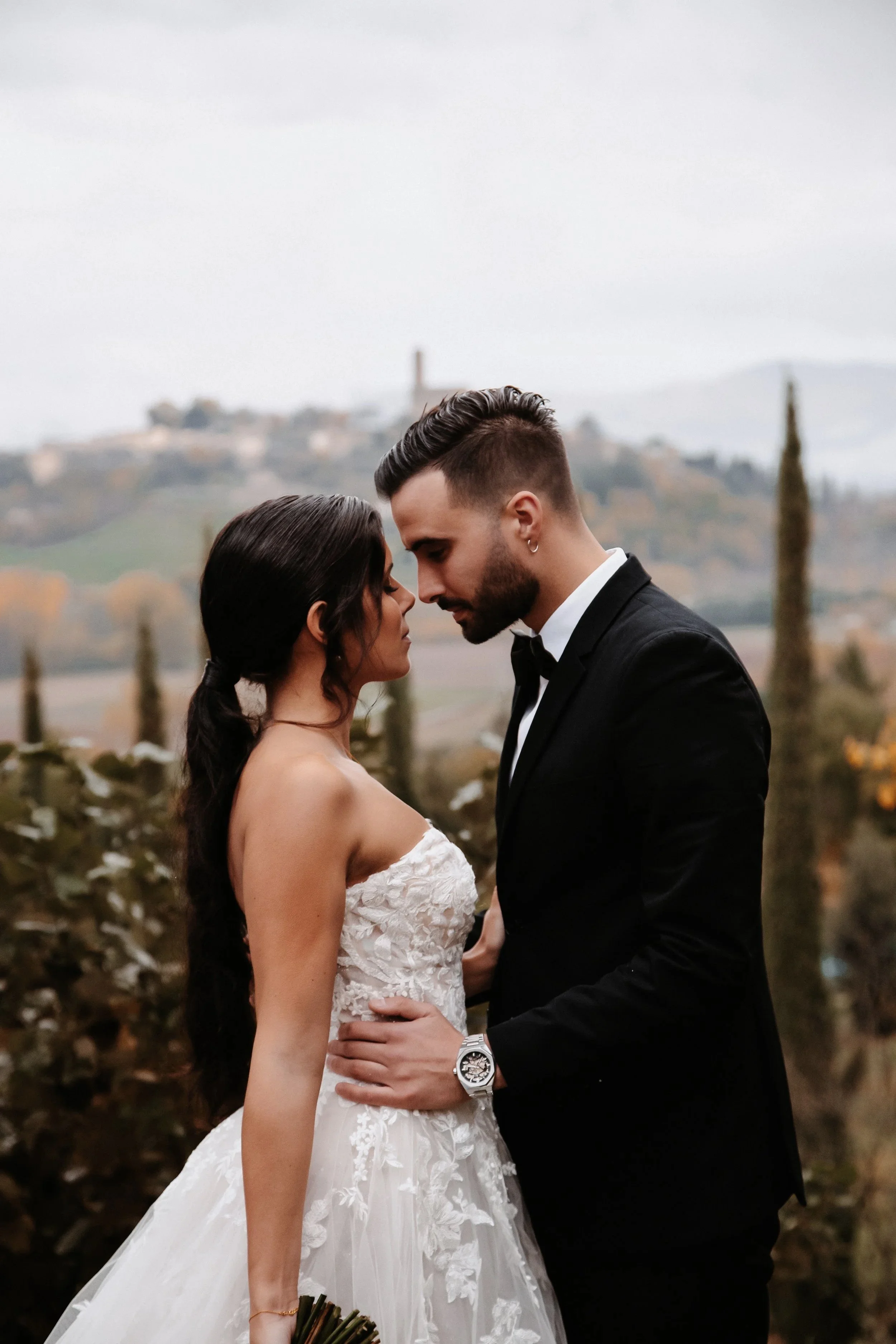 Ein Hochzeitspaar in eleganter Kleidung, die sich nahe stehen, mit einer landschaftlichen Kulisse im Hintergrund.
