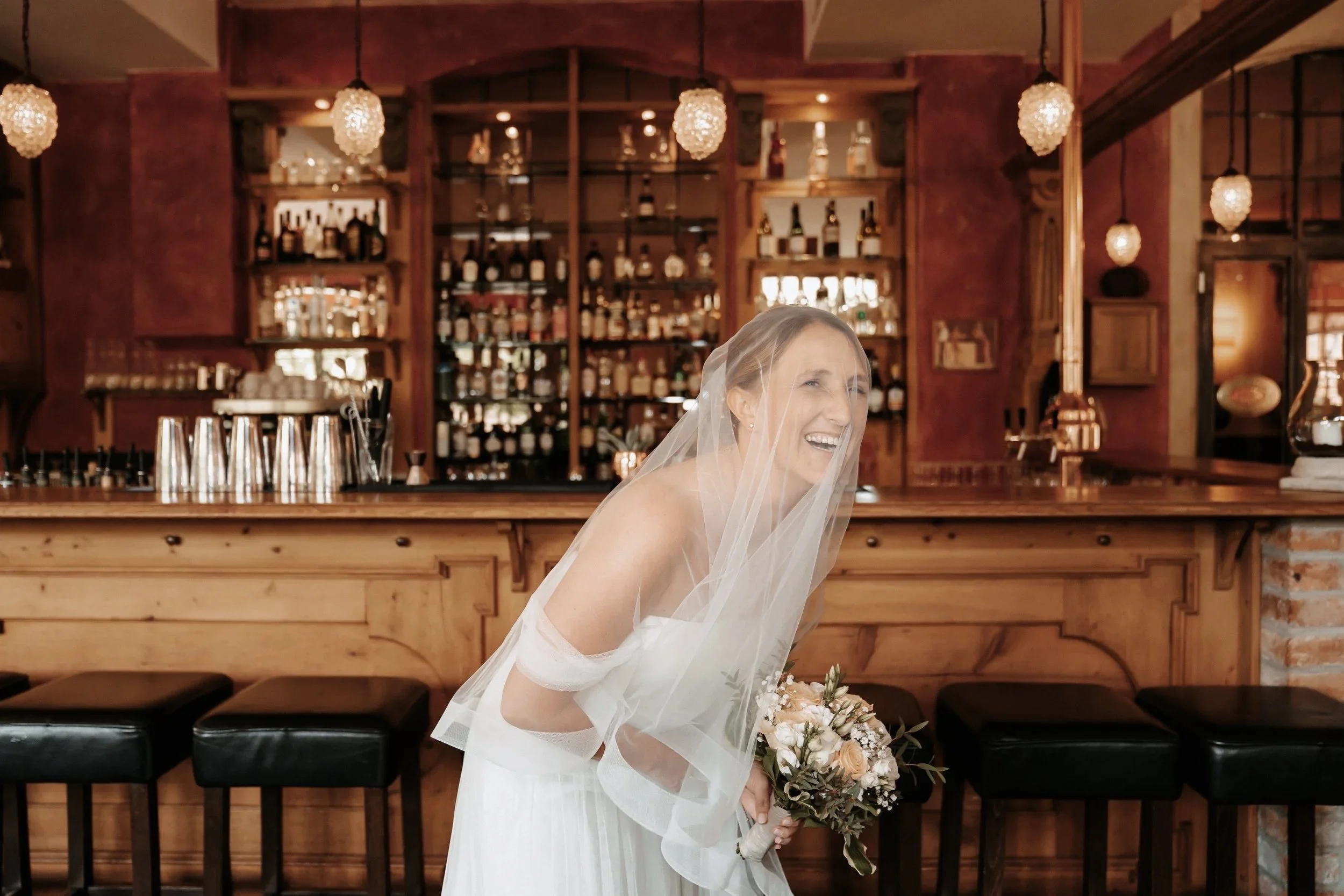 Eine Braut in einem weißen Kleid und Schleier lacht und hält einen Blumenstrauß. Im Hintergrund ist eine gut bestückte Bar mit Holzverkleidung und mehreren Getränkeflaschen zu sehen.