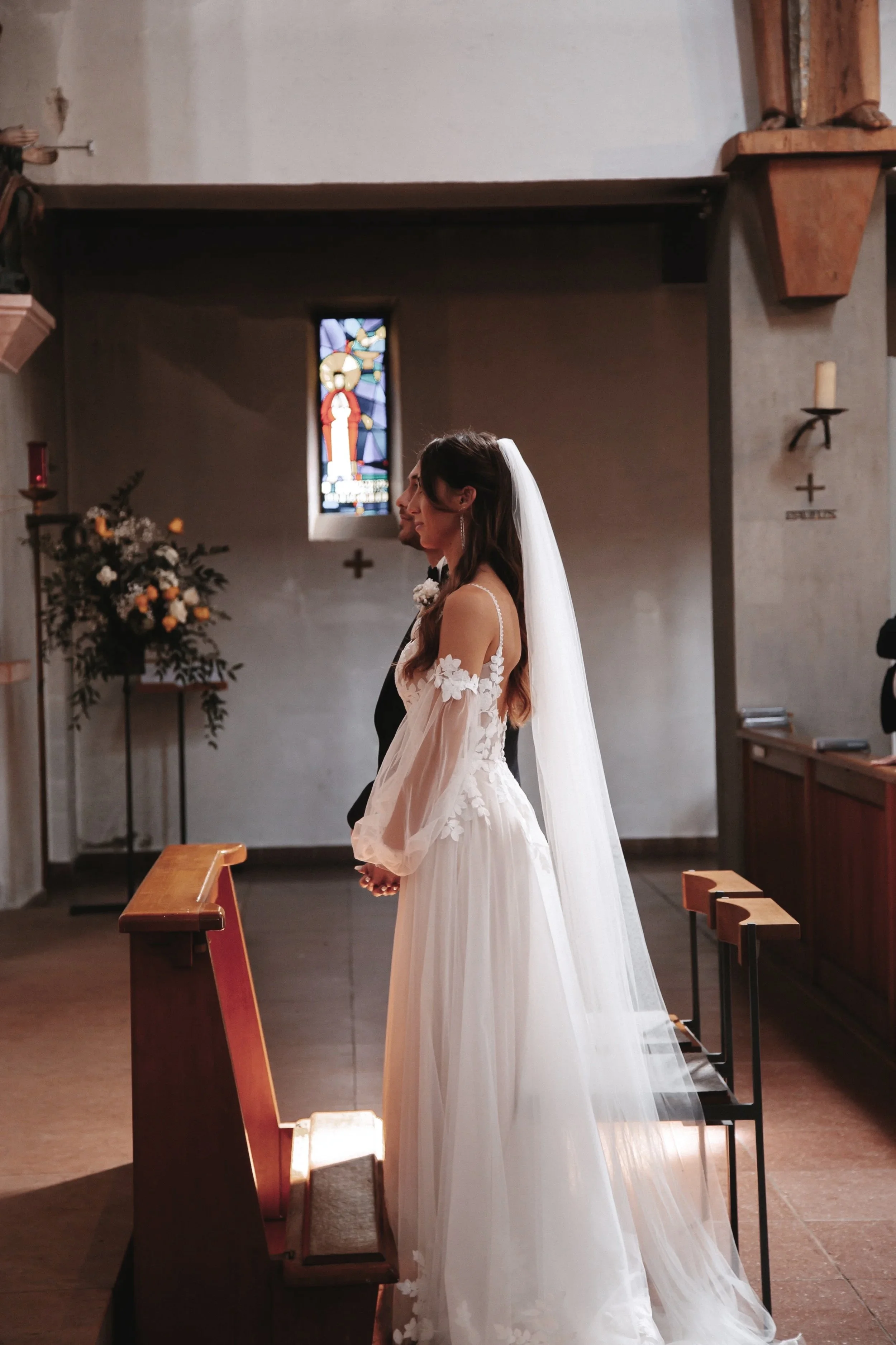 Brautpaar in einer Kirche bei einer Trauung, die Braut trägt ein langes weißes Kleid mit Schleier, im Hintergrund ein buntes Glasfenster und Blumenschmuck.